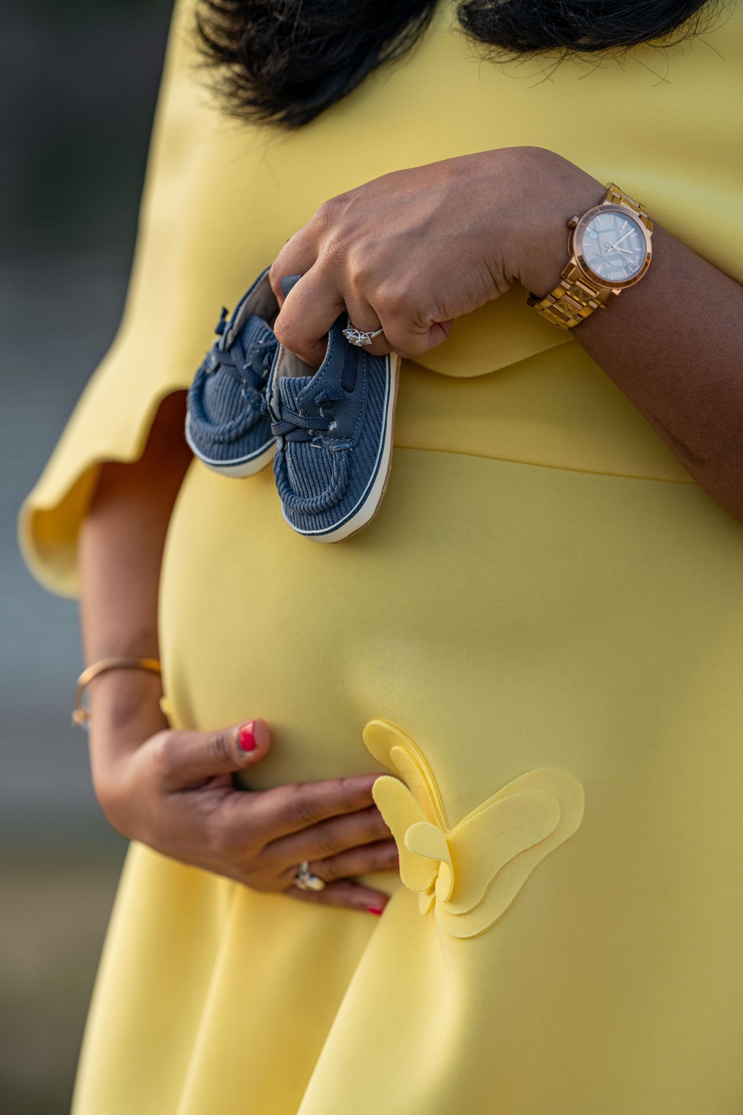 Pregnant woman in yellow dress holding tiny navy blue baby shoes over her baby bump, wearing a gold watch.