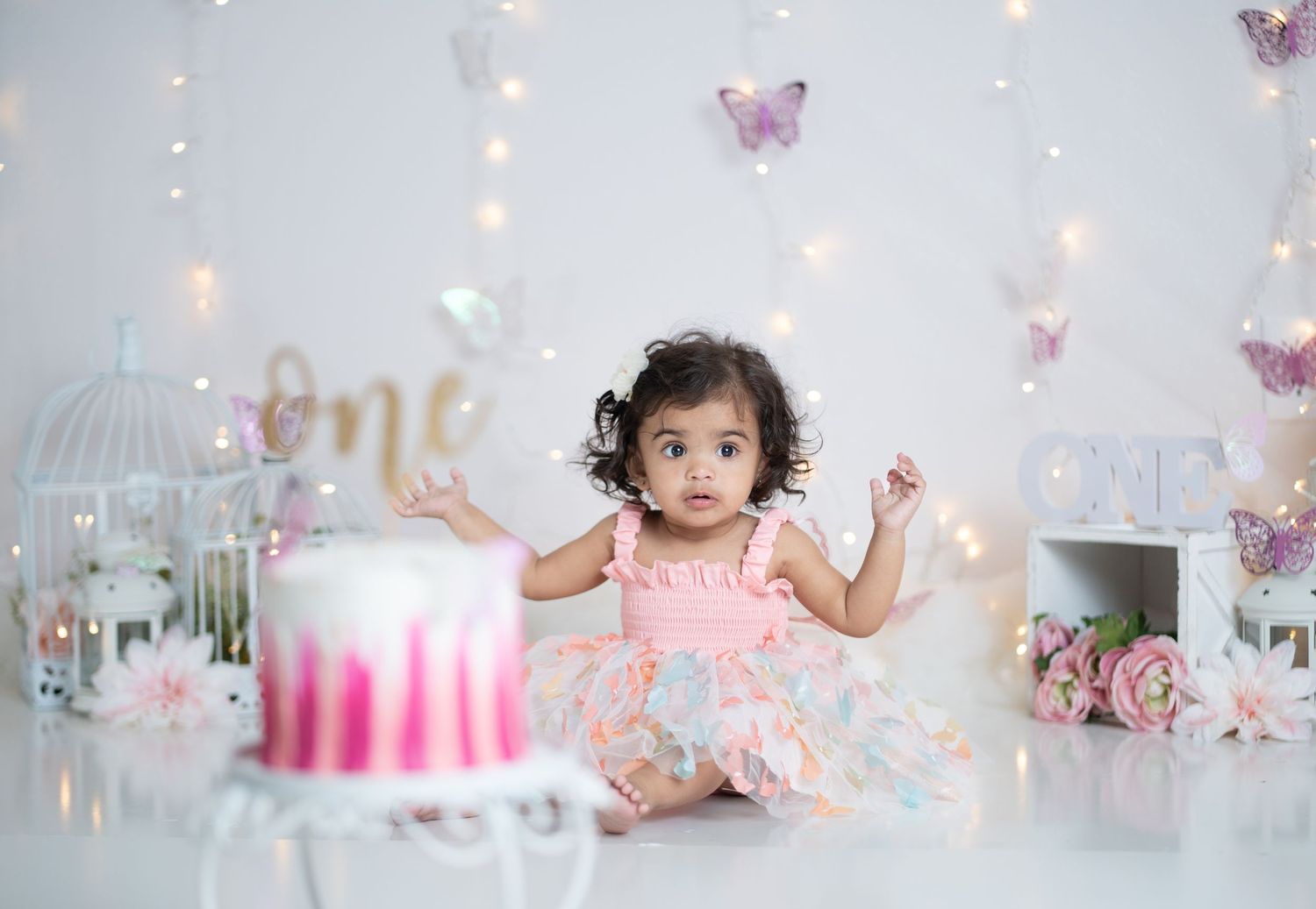 Baby girl in pink dress sits beside a first birthday cake with butterfly decorations and fairy lights backdrop.