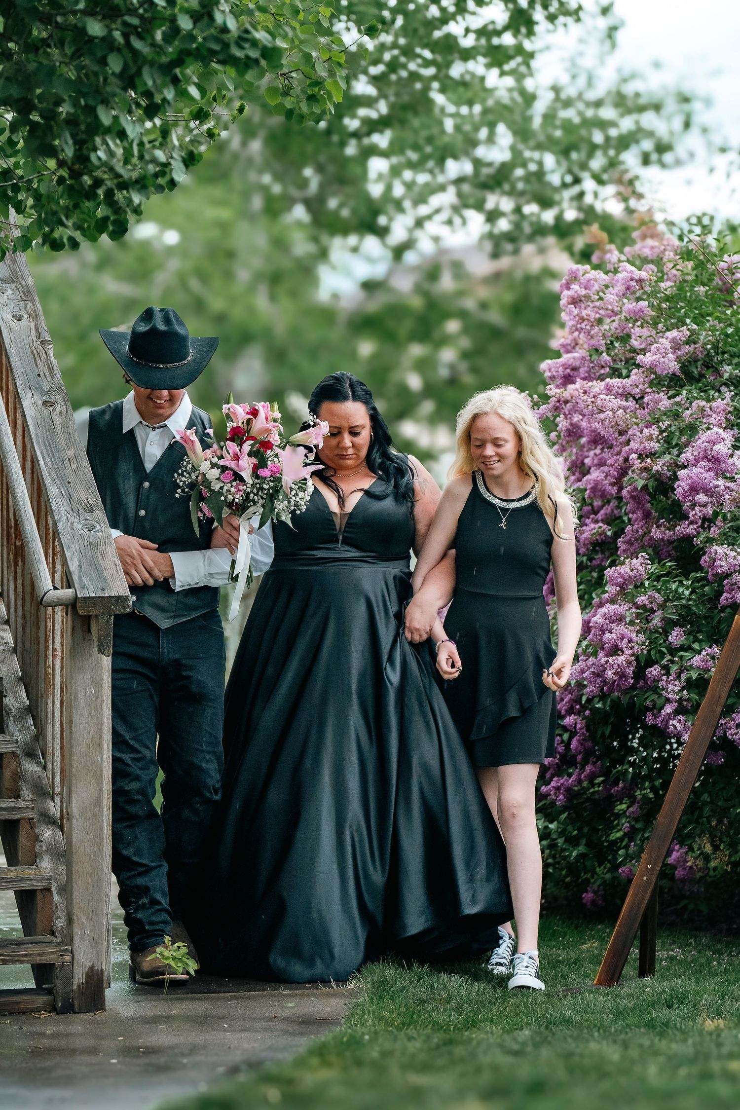A wedding party in formal attire walks down wooden steps surrounded by blooming lilac bushes.