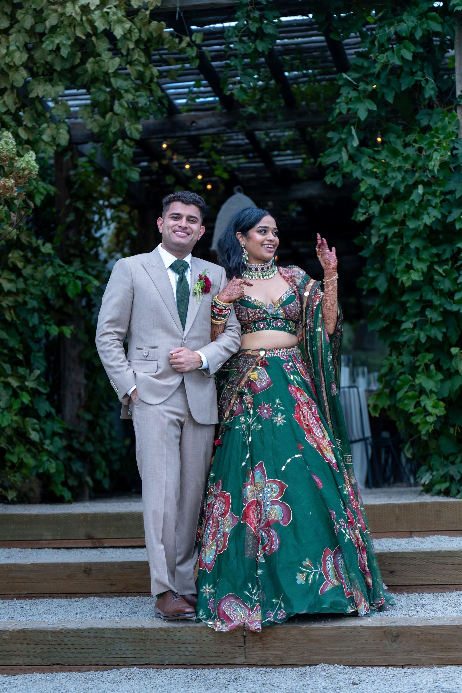 Wedding couple poses on garden steps with the bride wearing an elegant green floral lehenga and groom in beige suit.