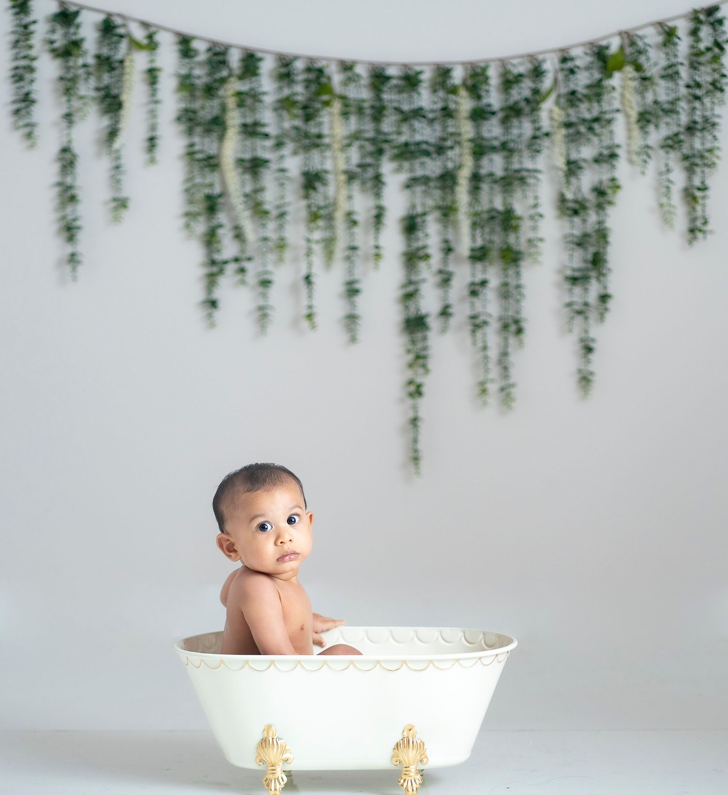 Baby sitting in decorative white bathtub with hanging greenery backdrop.