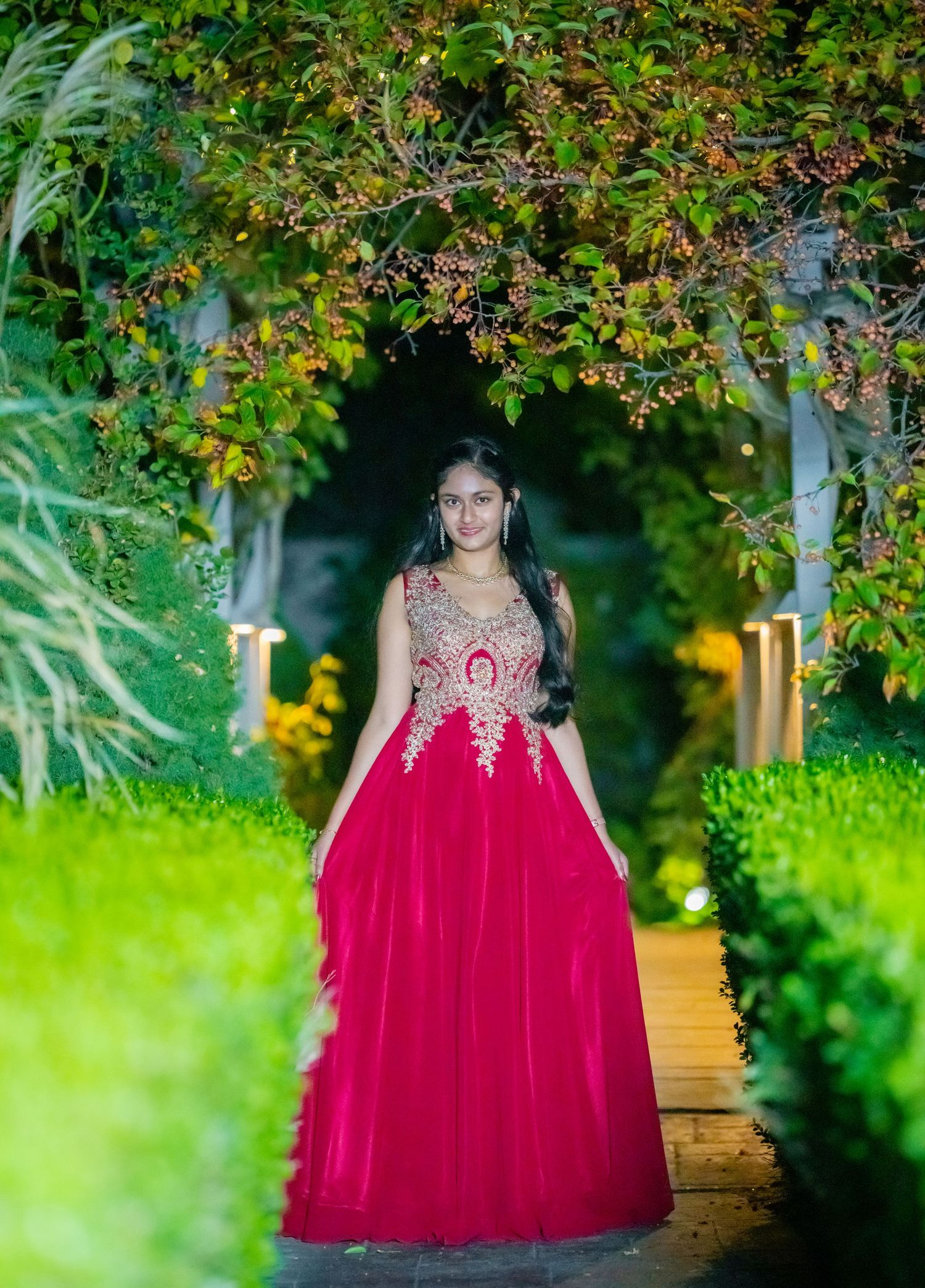 Formal portrait in red and gold embellished gown on garden pathway.