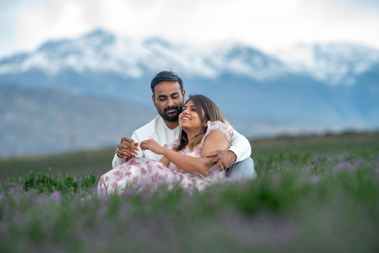 Couple embracing in mountain meadow with snow-capped peaks in background.