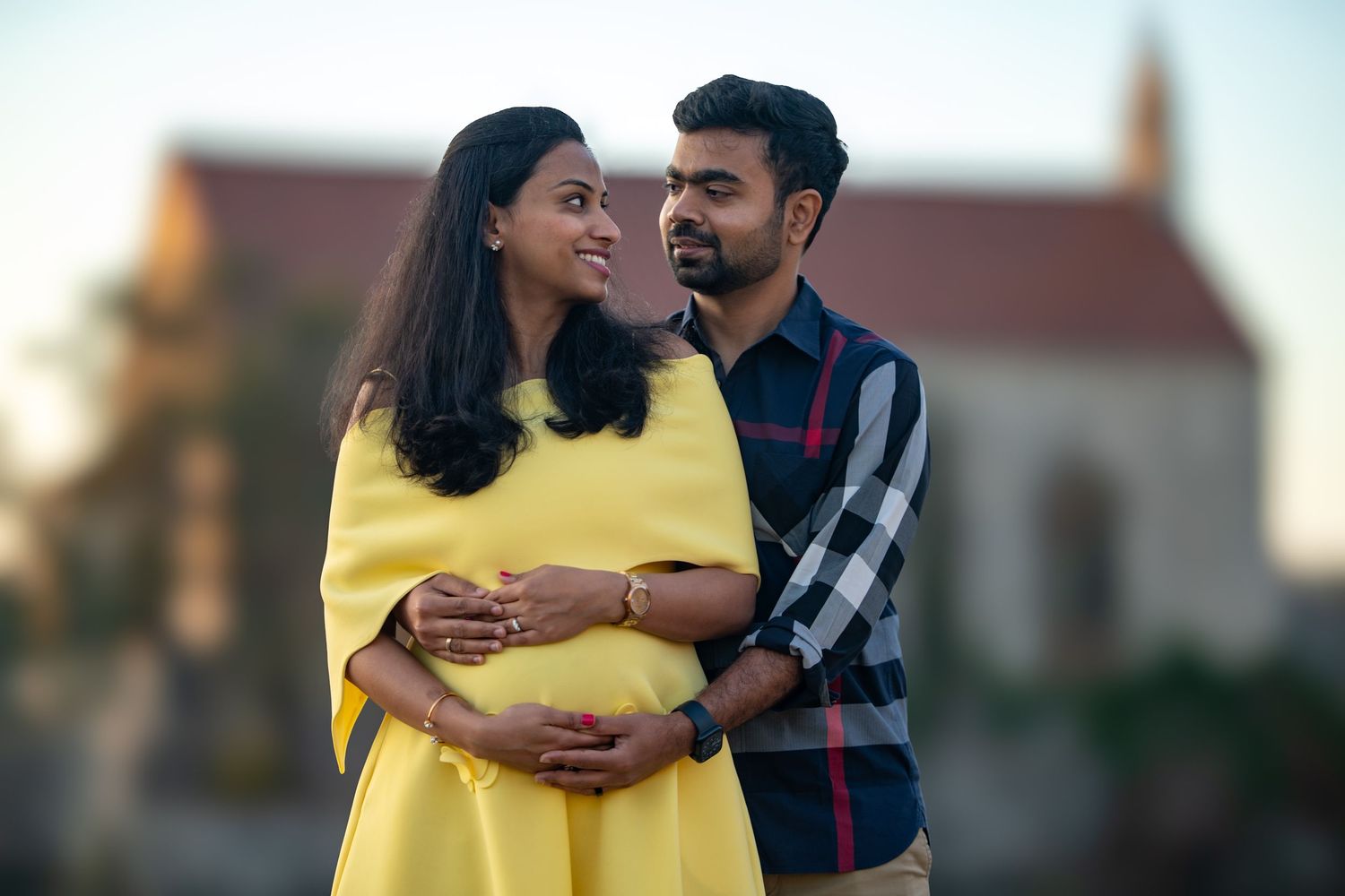 A pregnant woman in a yellow dress and her partner share a tender moment outdoors near a historic church.