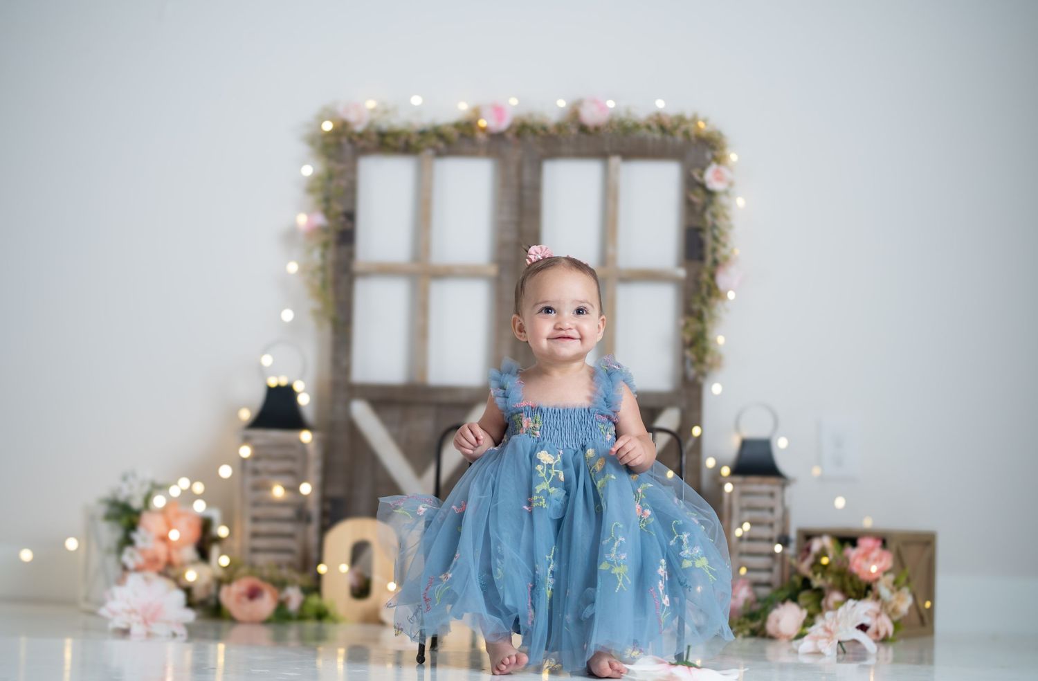 Baby girl in blue tulle dress smiles among fairy lights, flowers, and rustic decor in a professional portrait.