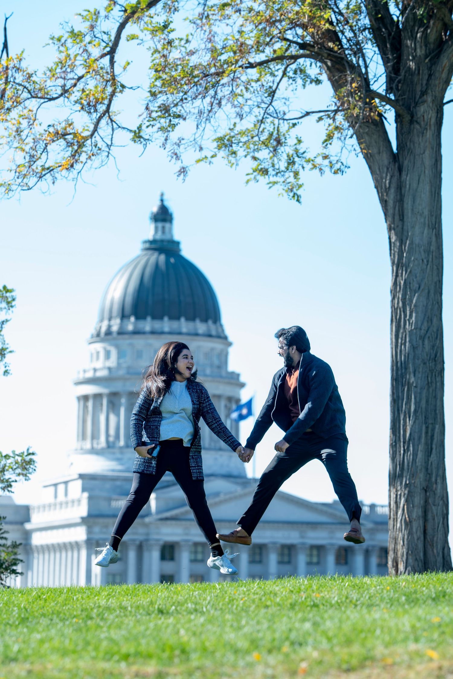 Playful couple jumping together in front of state capitol building on sunny day.