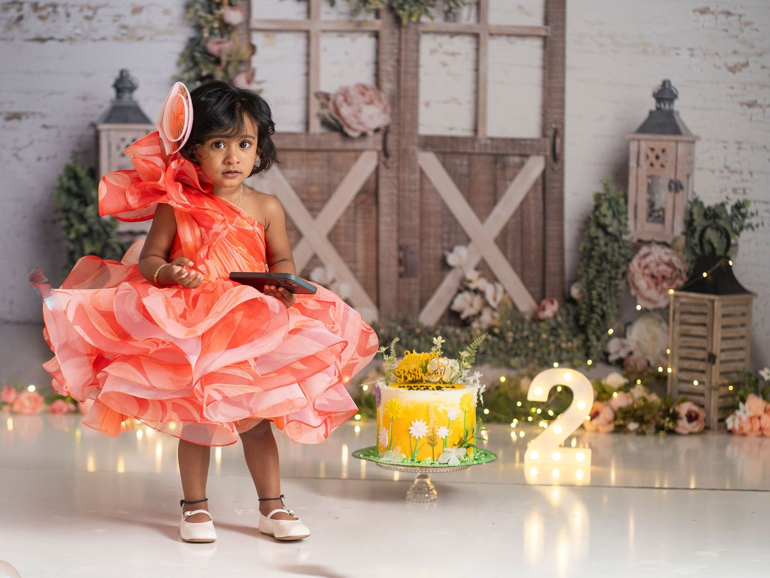 Toddler in coral ruffle dress celebrating second birthday with rustic barn door backdrop and twinkling lights.