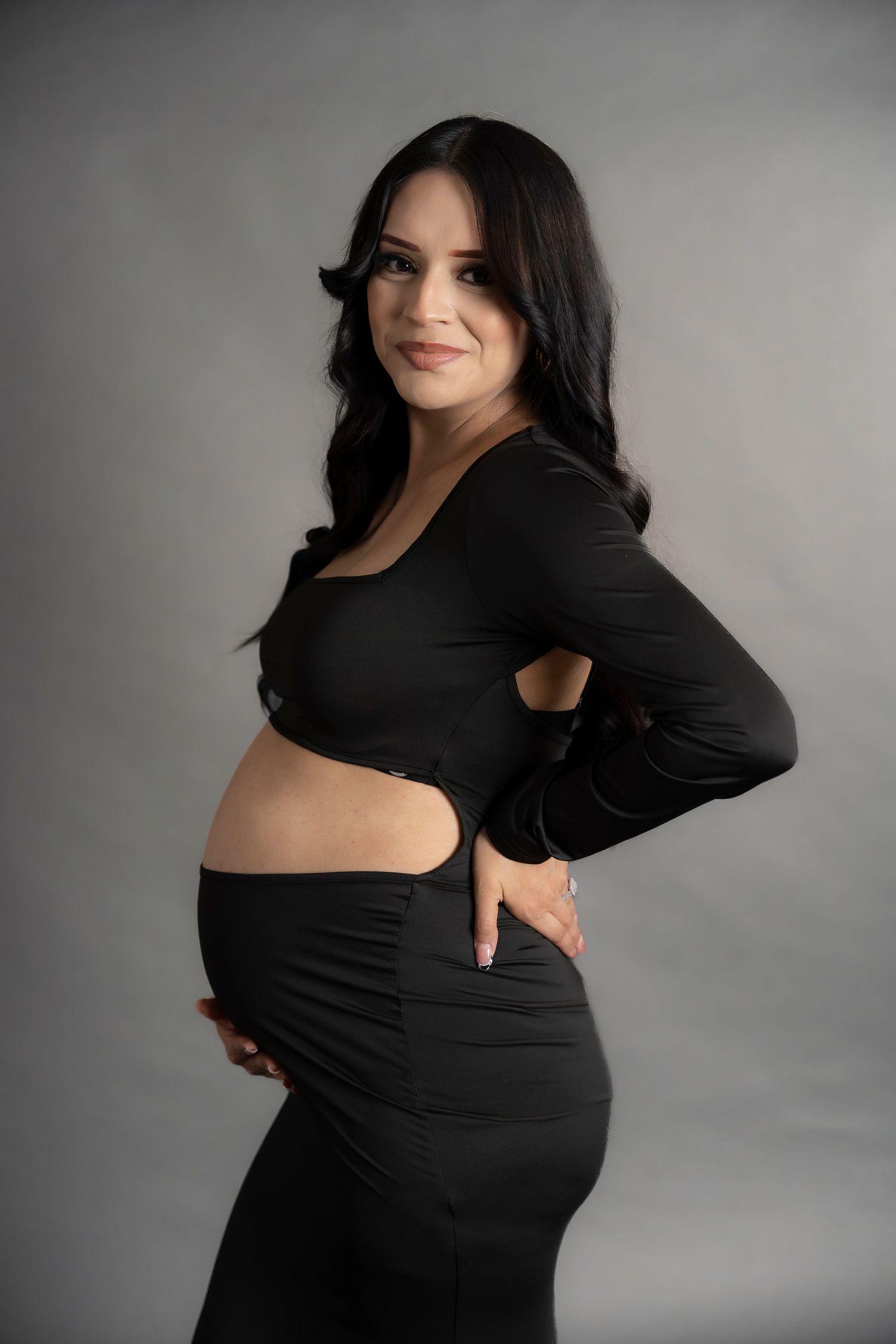 Maternity portrait of woman in black dress with cutout details against gray backdrop.
