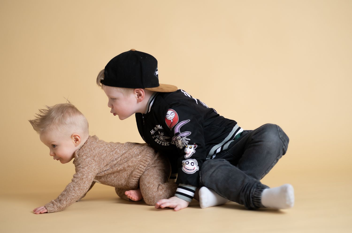 Two children playing together on a beige backdrop, one crawling while wearing a brown sweater.