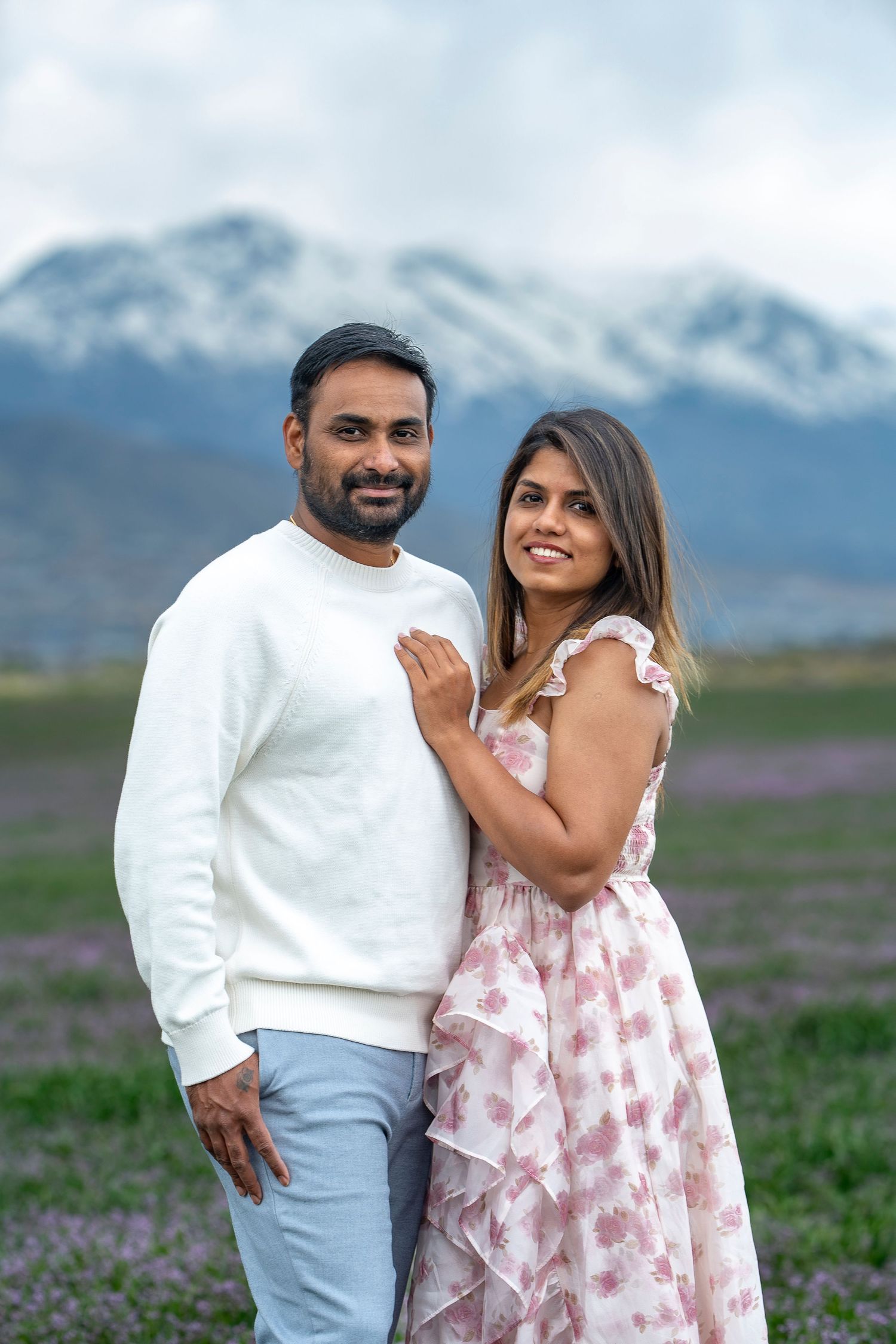 Engaged couple poses in mountain field wearing white and pink spring outfits.