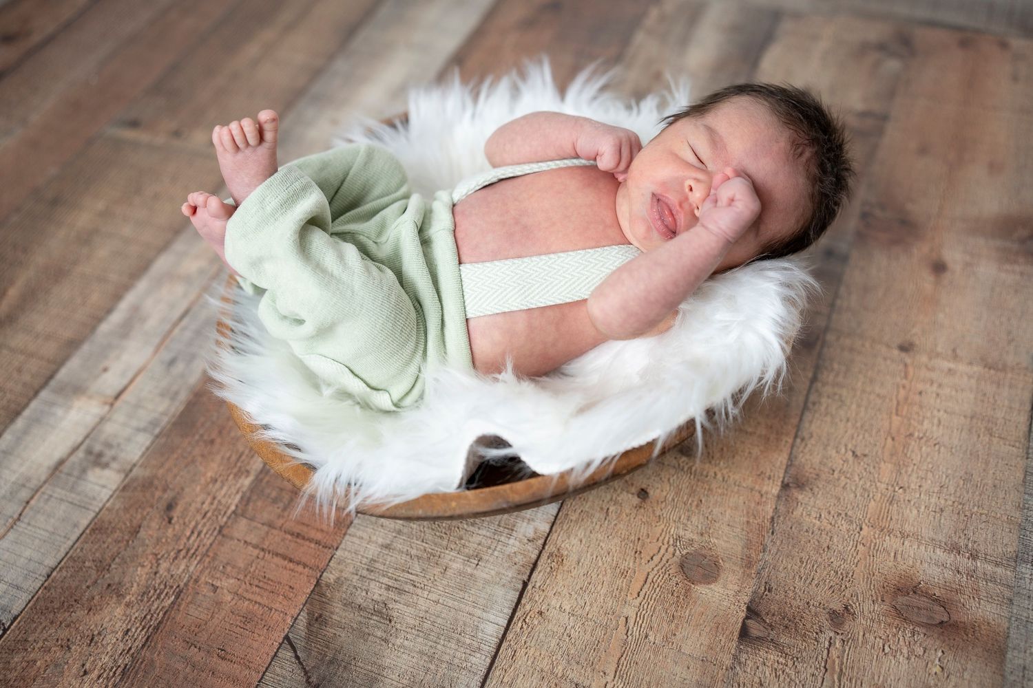 Sleeping newborn on white fur wearing mint green outfit on wooden floor.