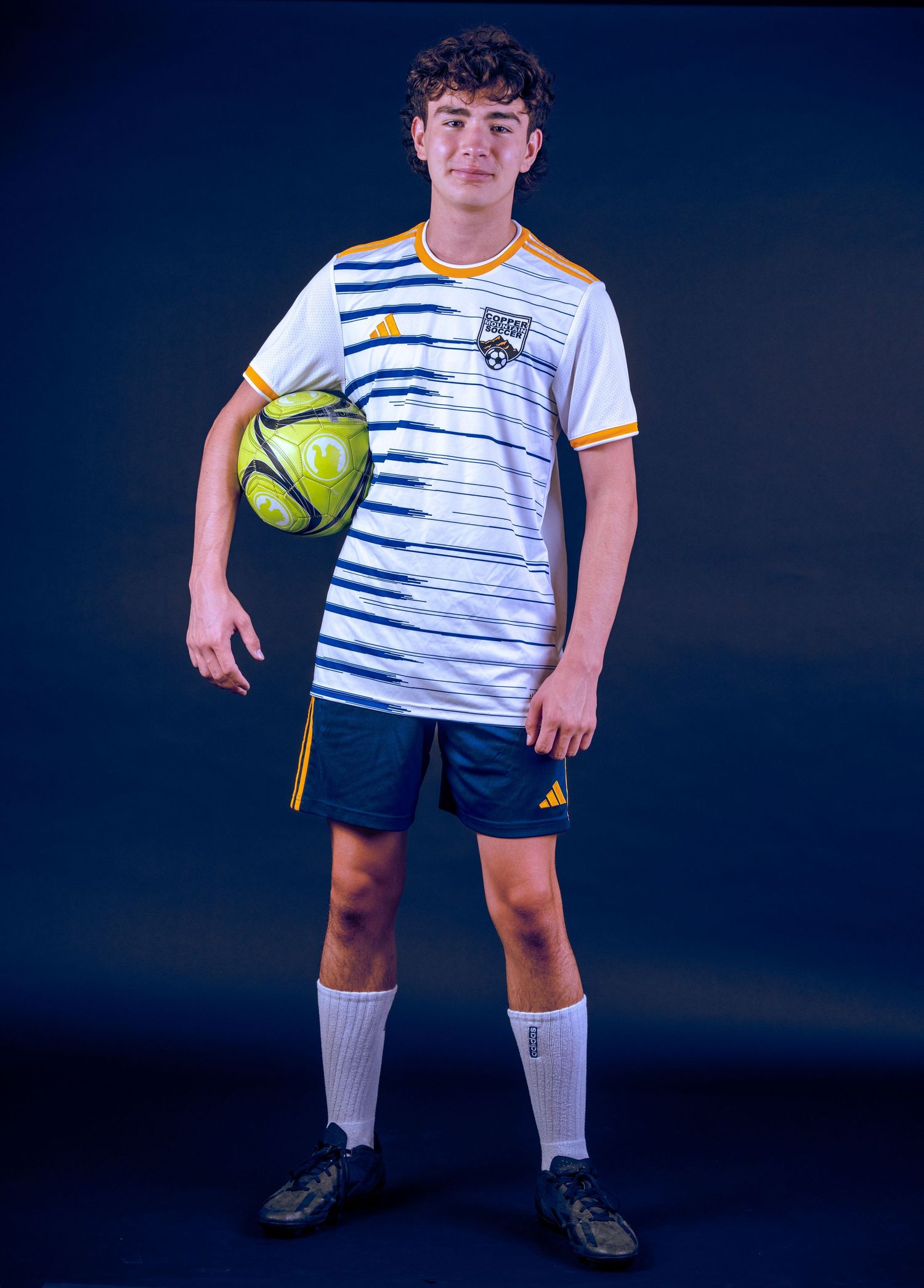 Young athlete in striped soccer uniform holds yellow ball against blue background.