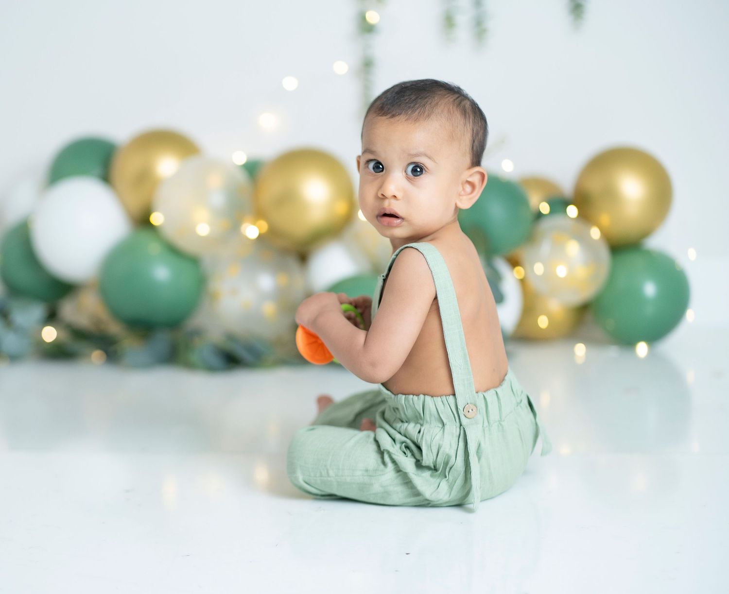 Baby in mint green outfit sitting against balloon arch backdrop.