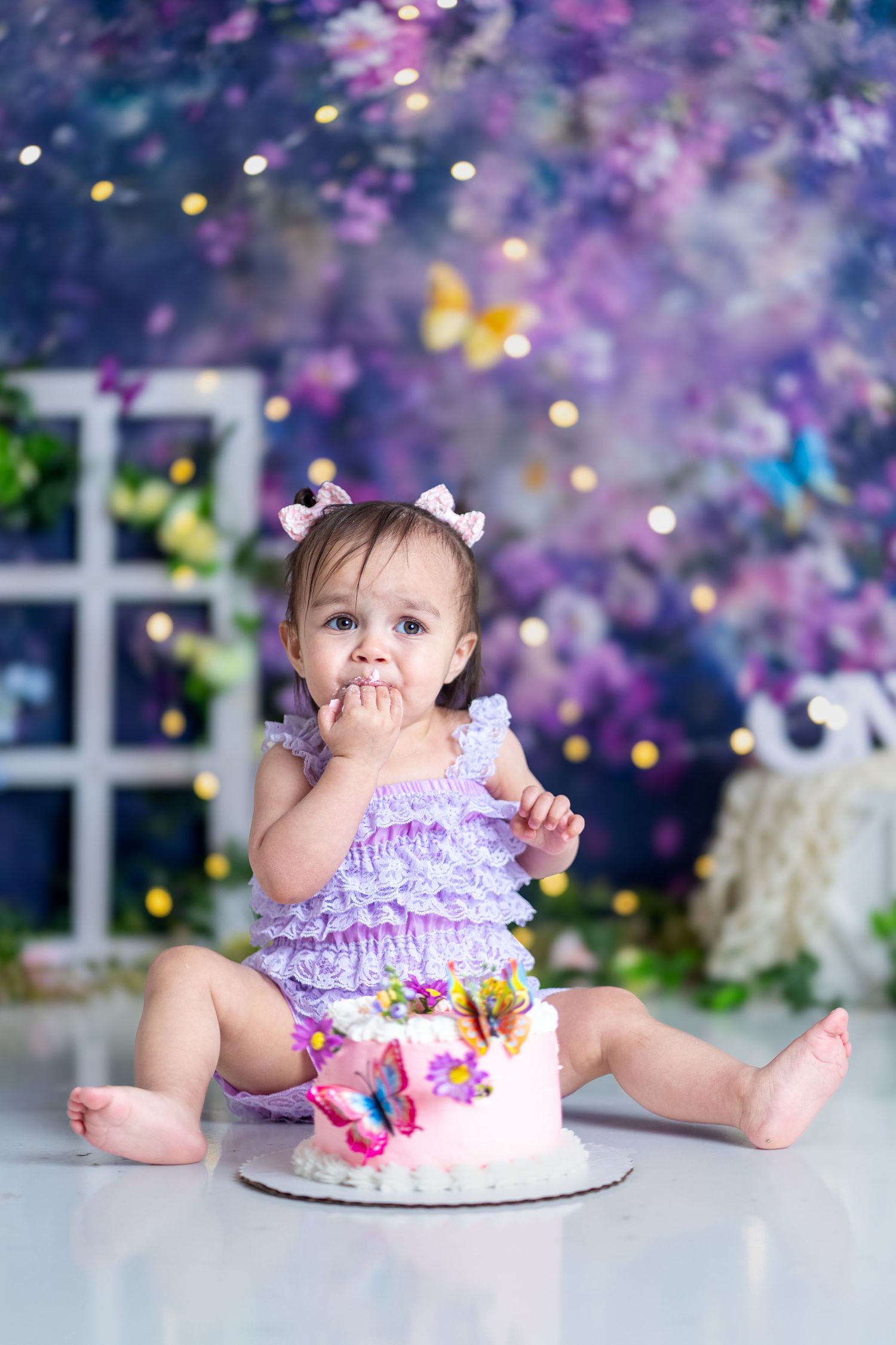 Purple romper cake smash session with magical bokeh lights and floral backdrop.