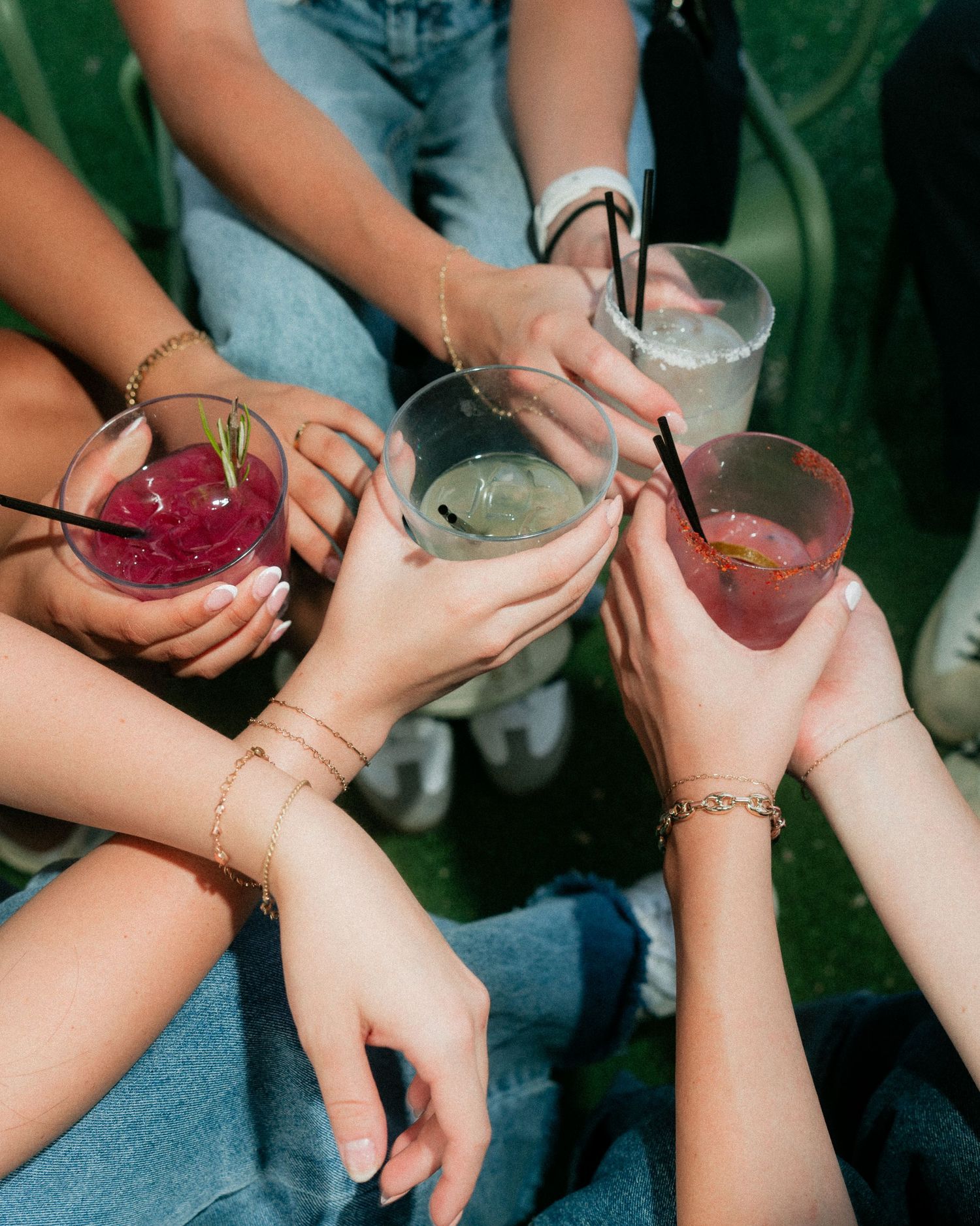 Friends clinking colorful cocktail glasses together in sunny outdoor celebration.