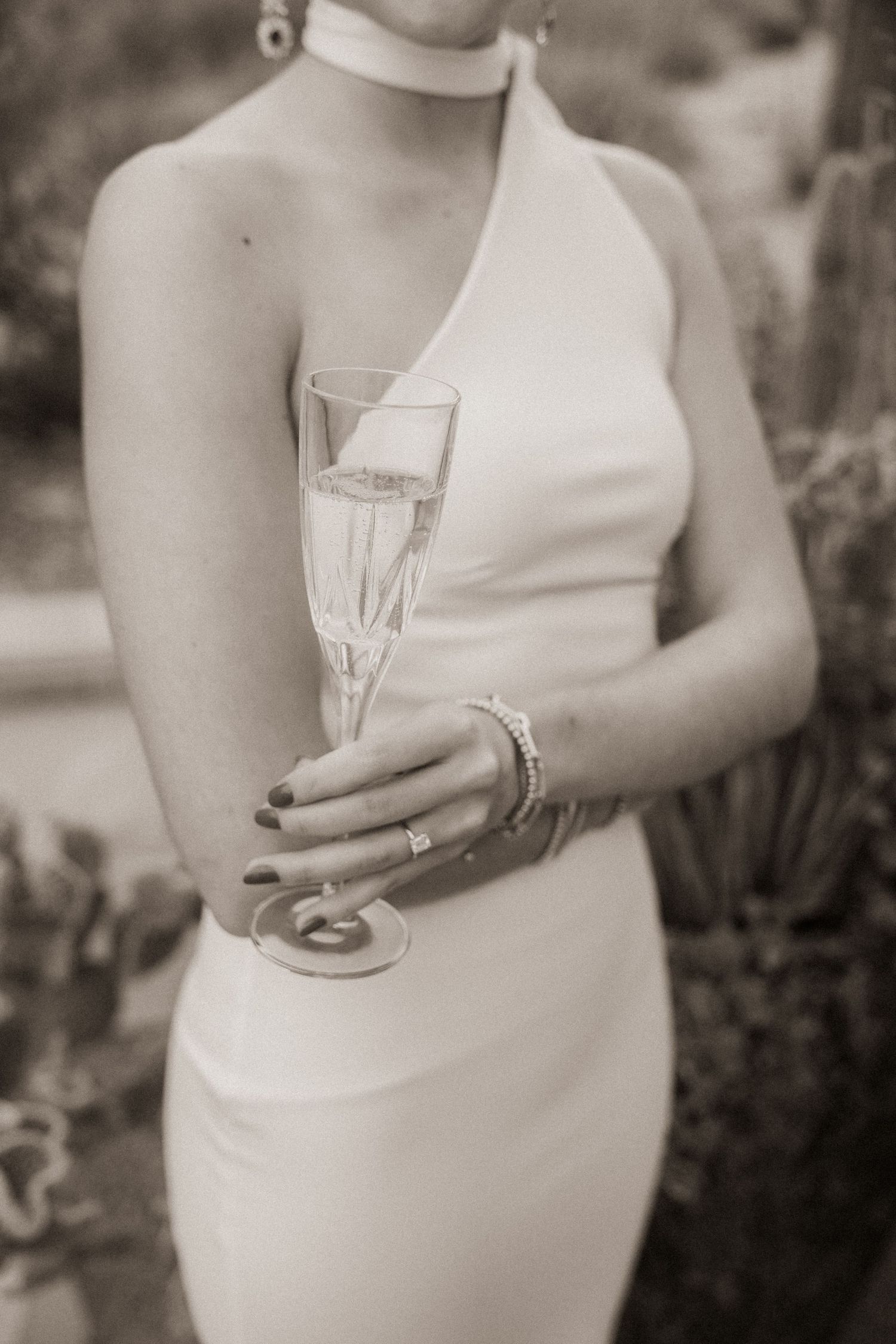 Bride in sleek white halter wedding dress holds a champagne flute in a sepia-toned photo.