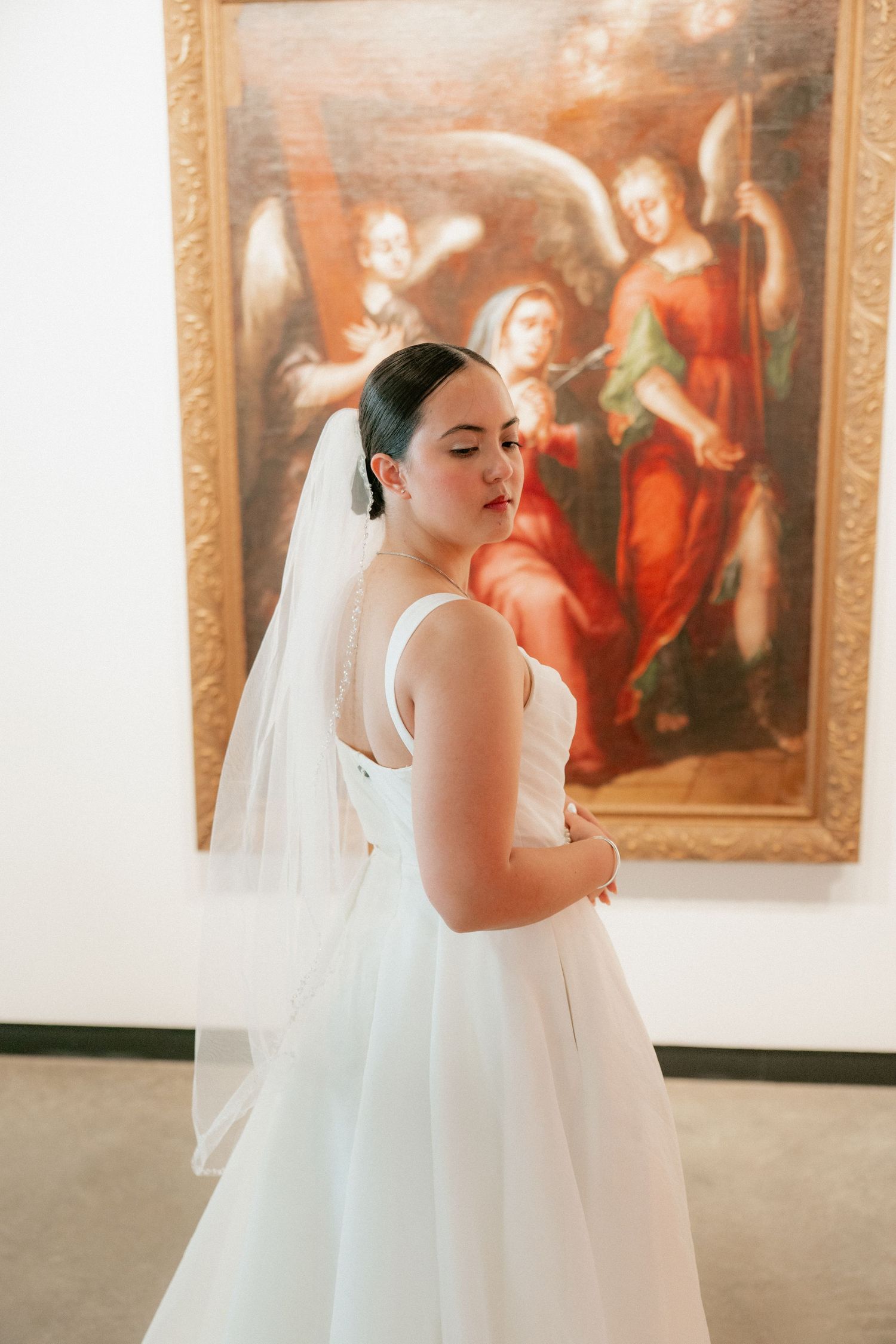 Bride in white gown and veil posing in front of a classical painting of angels in an art museum.