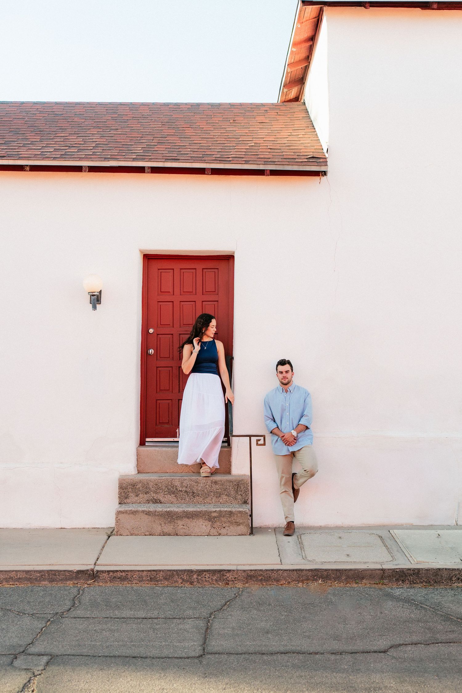 Engaged couple posing by a white building with a red door in a warm desert town setting.