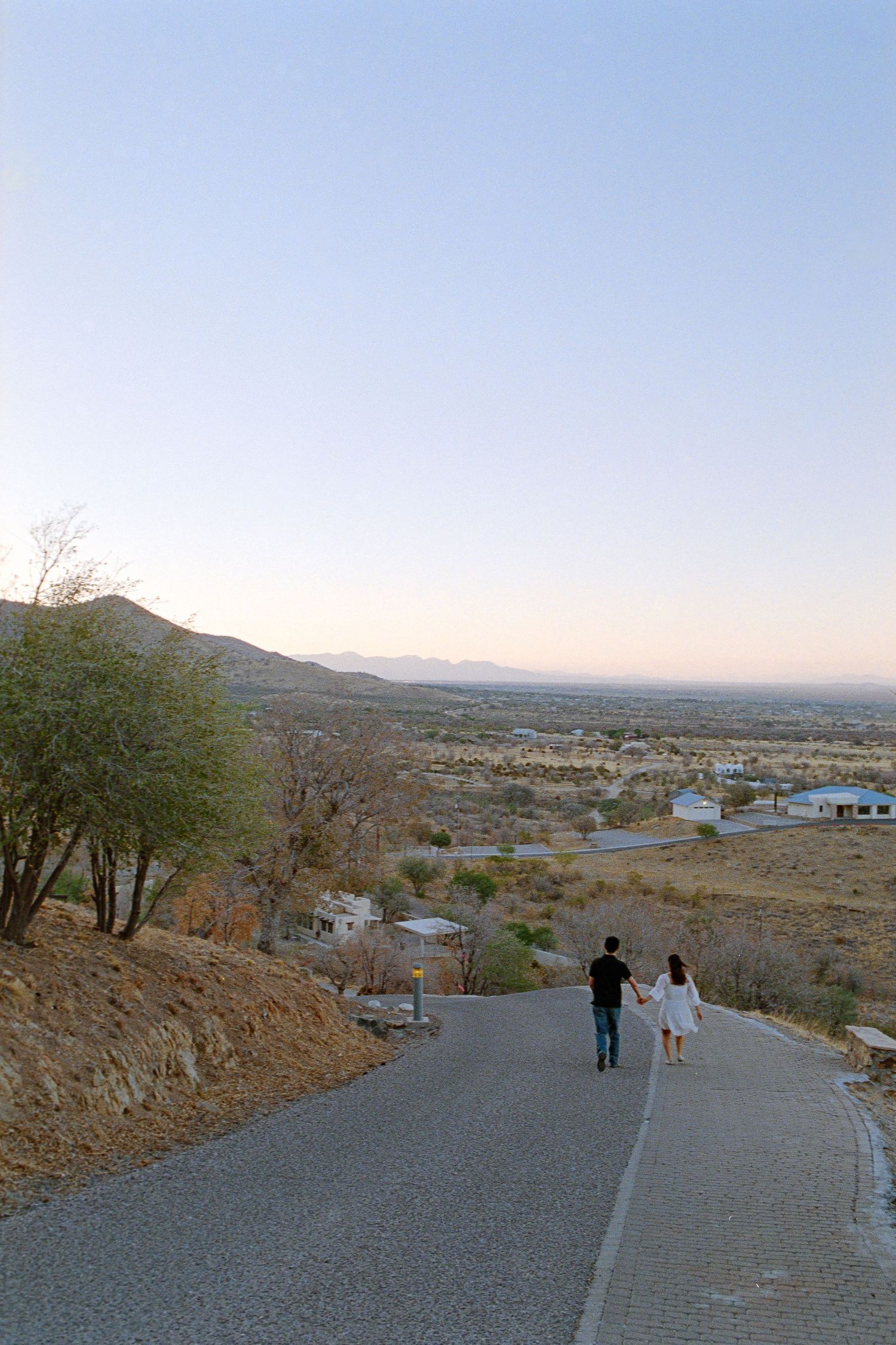 Couple walking hand in hand down a scenic desert road at sunset with mountain views.