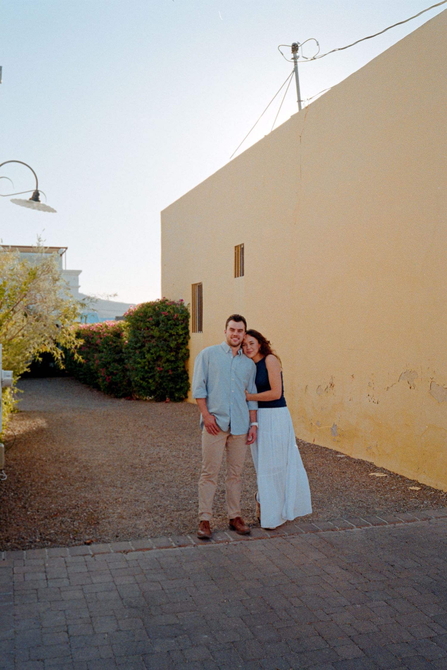 A couple poses together beside a yellow stucco wall in an outdoor courtyard setting.