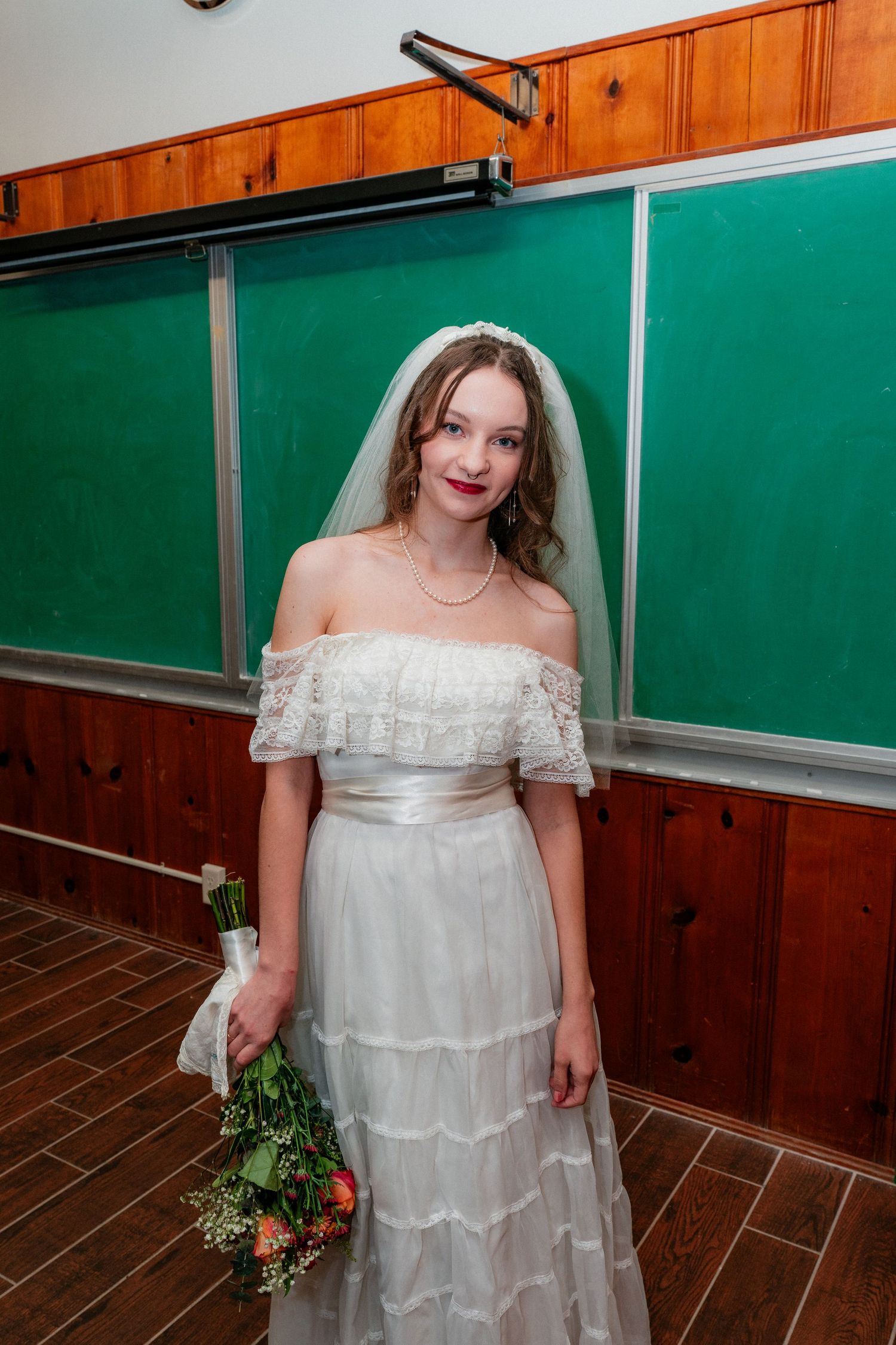 Elegant bride in off-shoulder white lace gown holding bouquet, standing before green chalkboards.