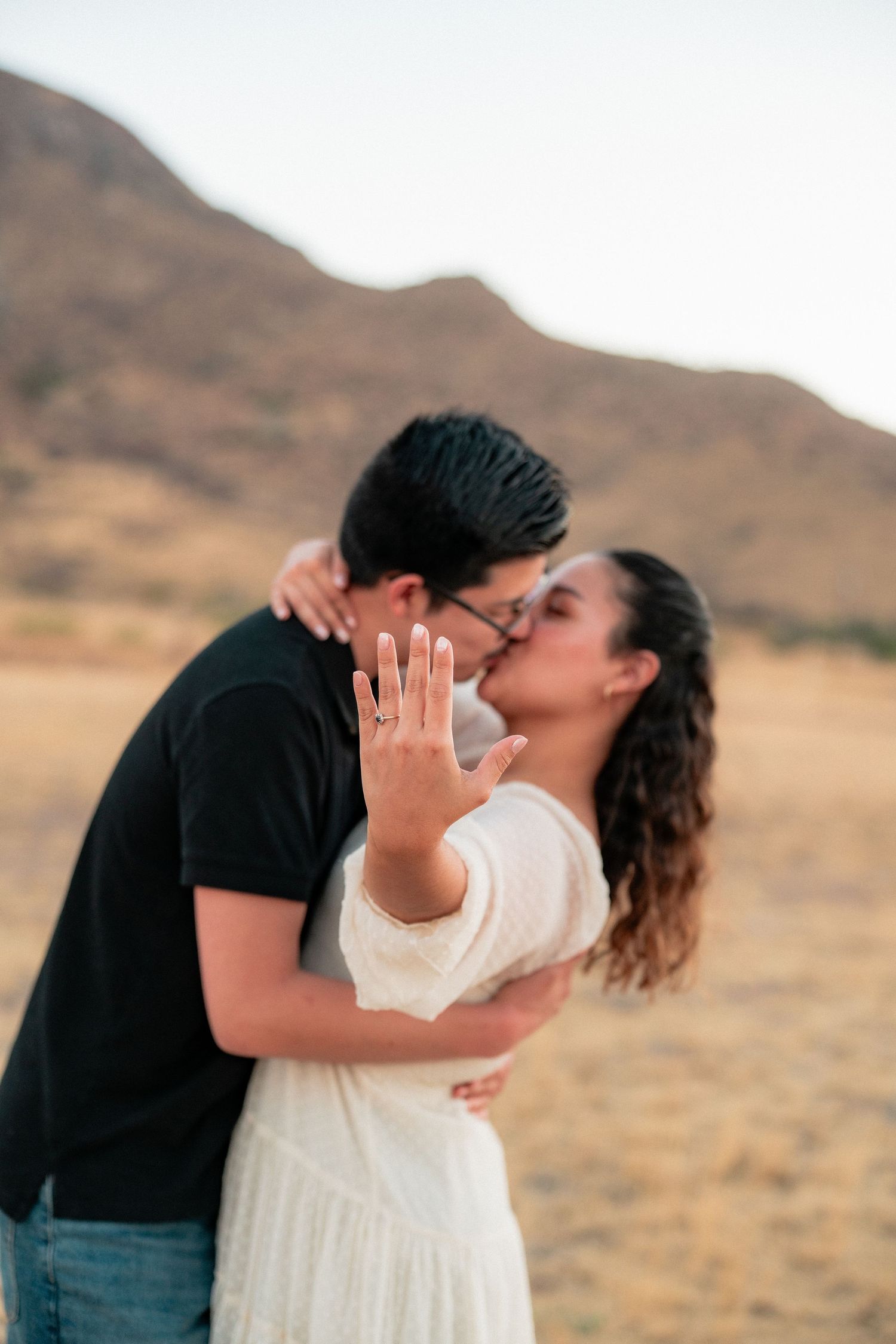 Newly engaged couple kissing while woman shows off her diamond engagement ring outdoors.