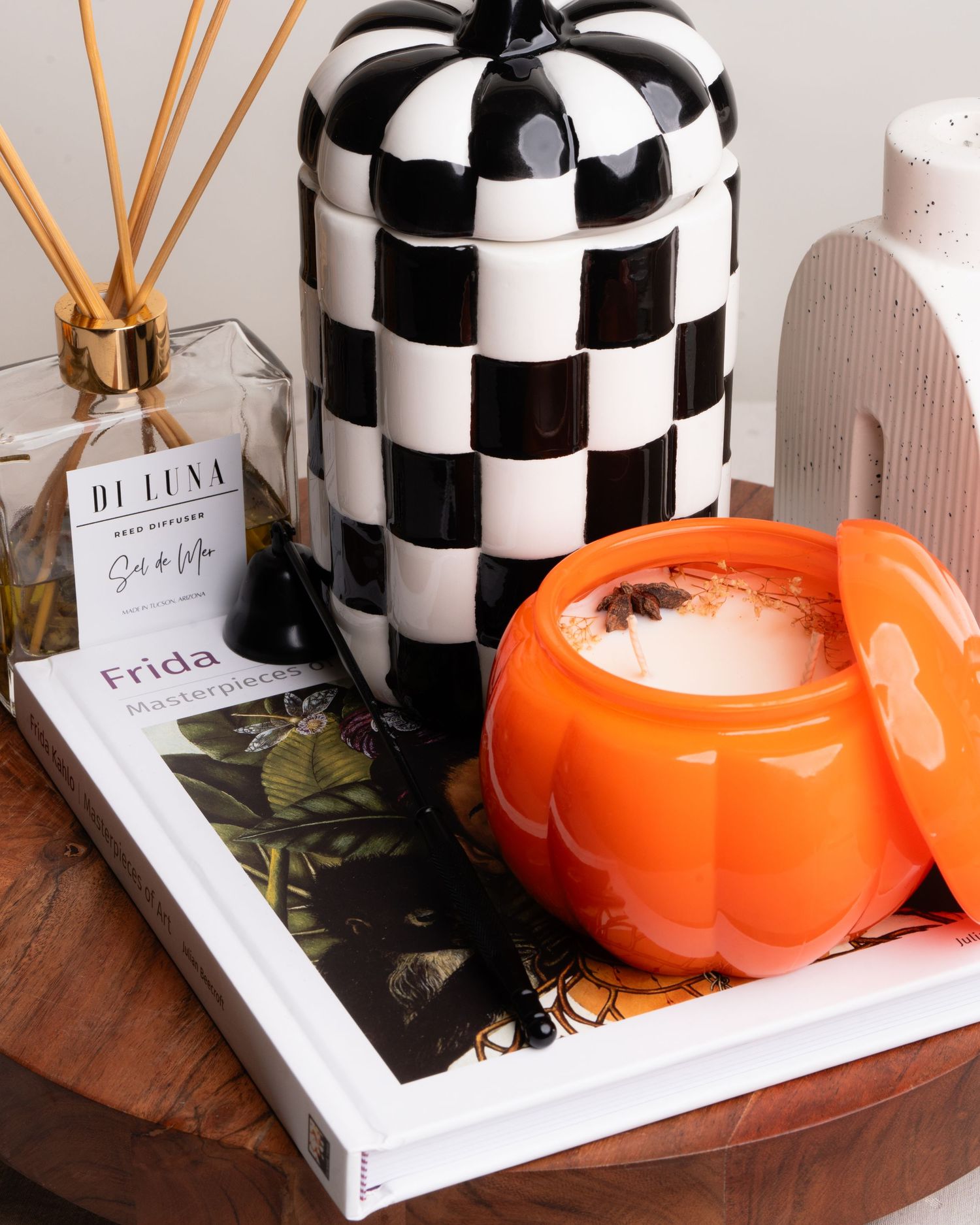 Orange pumpkin candle beside checkered ceramic jar with reed diffuser and Frida art book.