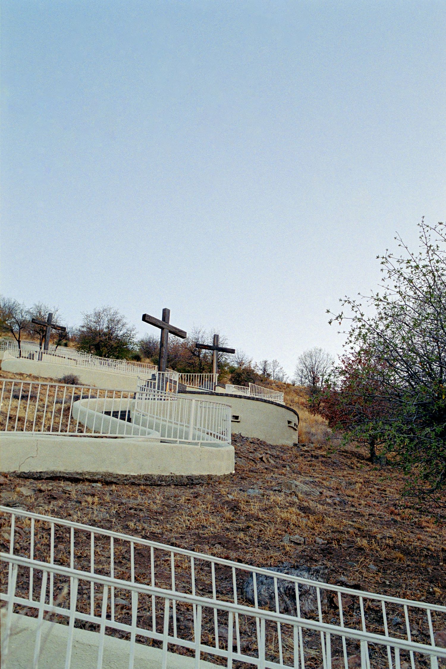Two wooden crosses on a hillside shrine with white fence railing under clear blue sky.