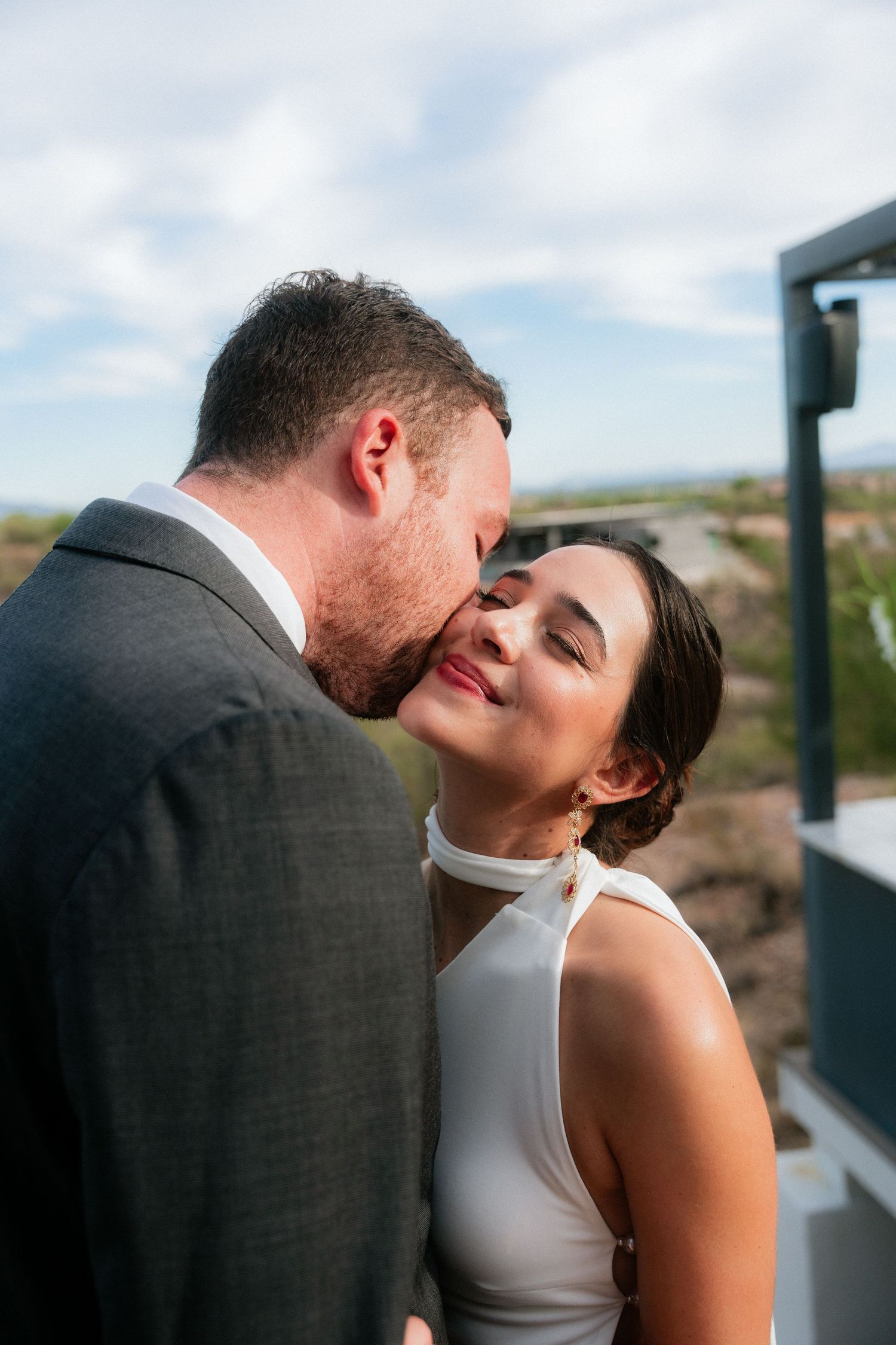 Groom tenderly kisses bride on the cheek outdoors against a cloudy sky backdrop.