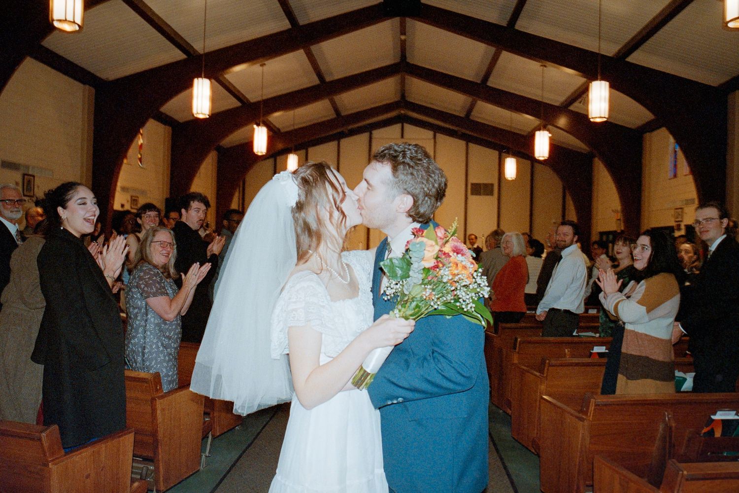 Newlyweds share romantic first kiss in church aisle as wedding guests celebrate around them.