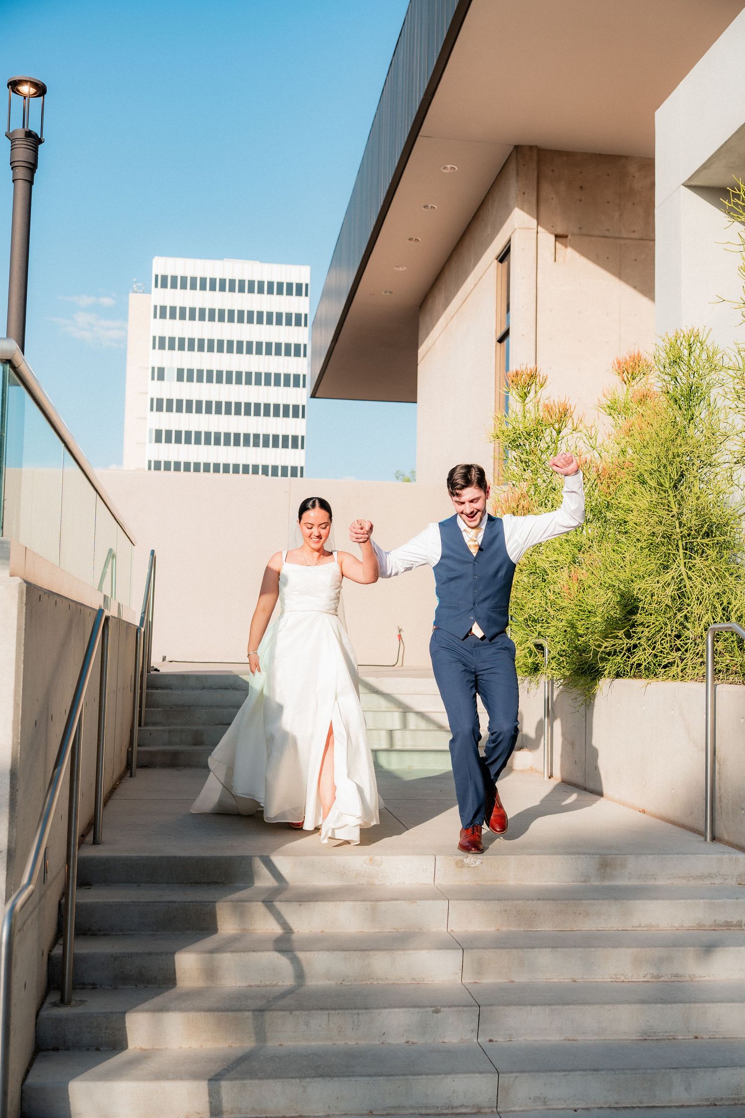 Joyful bride and groom descend outdoor steps hand-in-hand at a sunny urban wedding venue.
