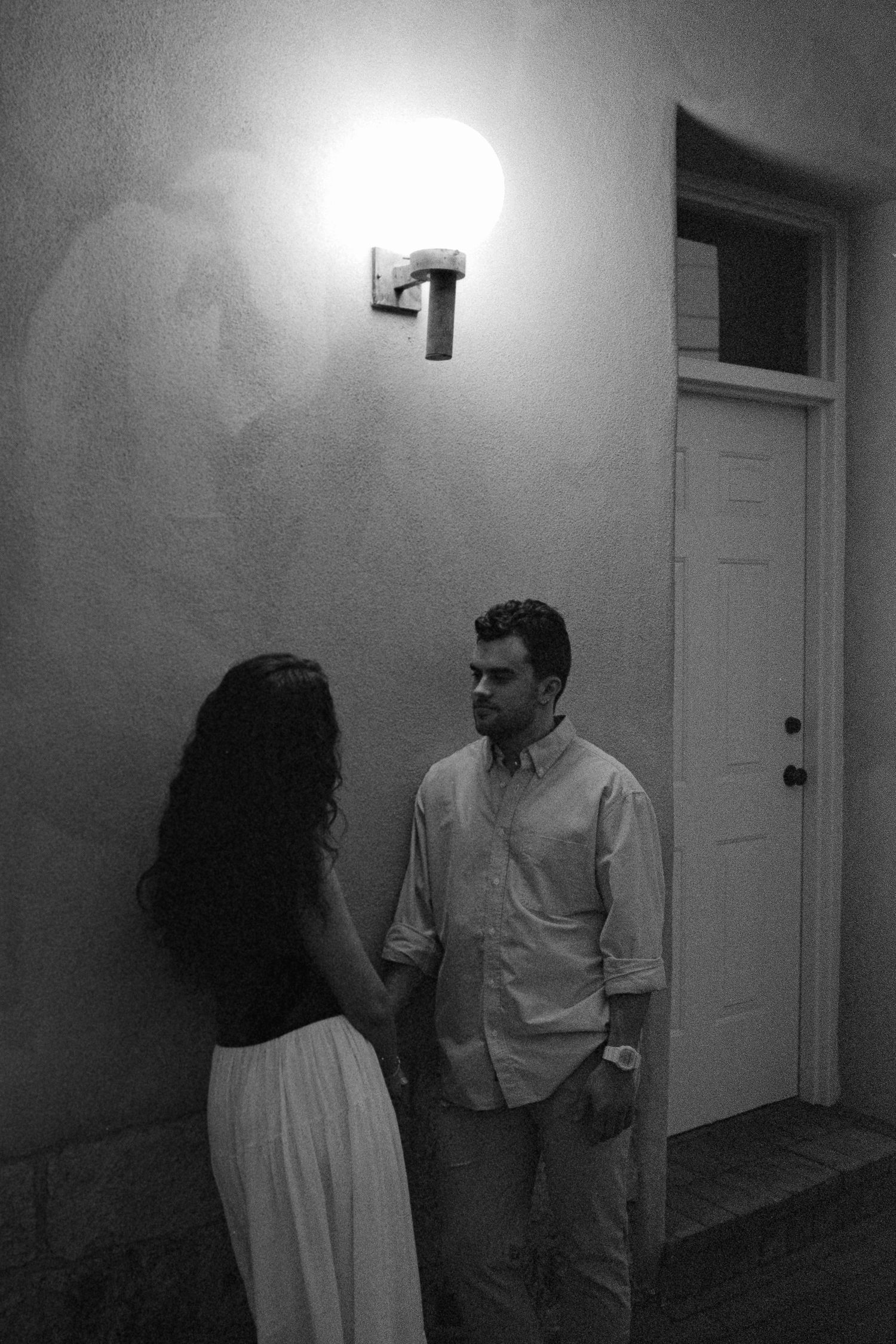 Black and white photo of couple holding hands under a glowing wall light at night.