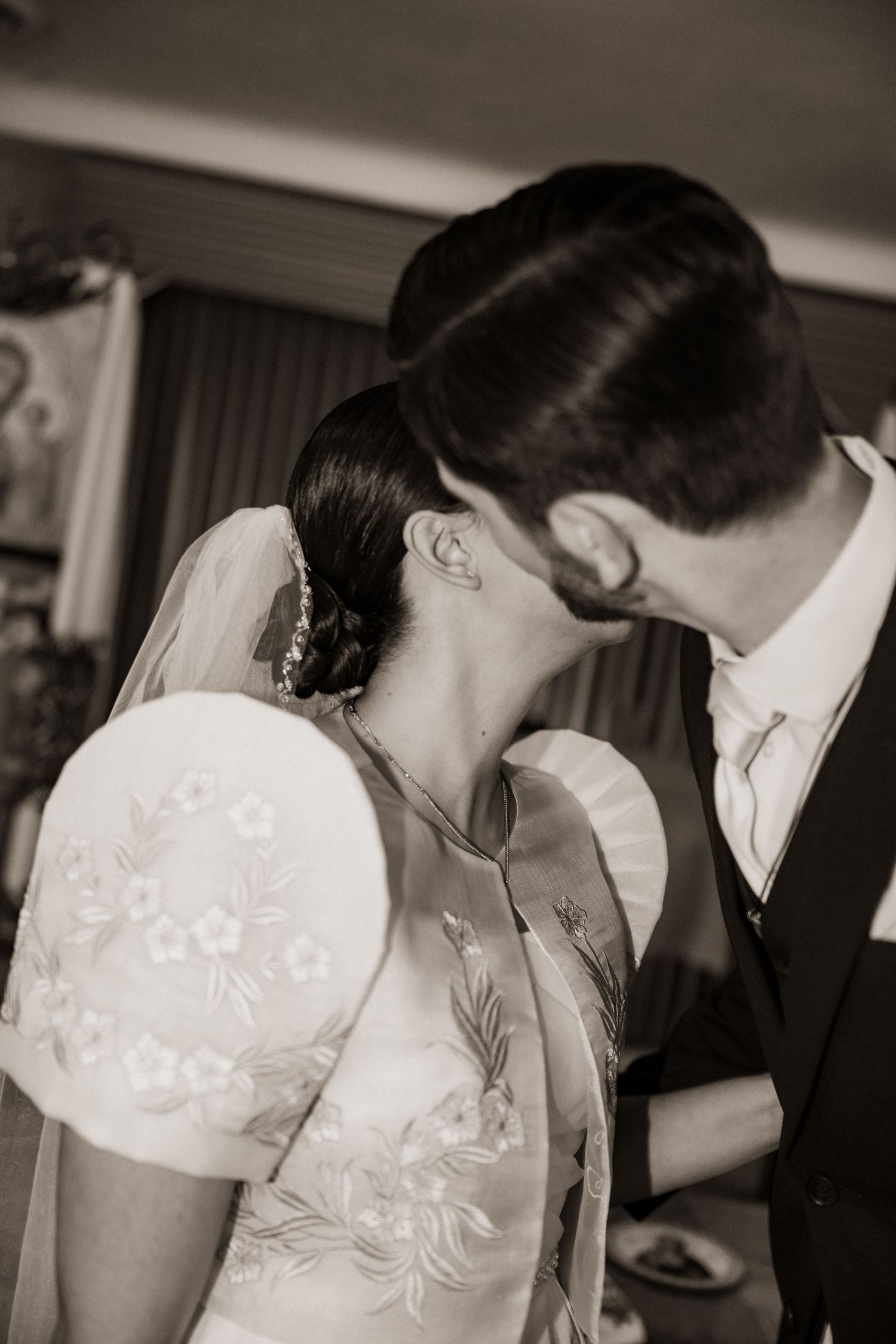 Black and white close-up of bride and groom about to kiss, bride wearing veil and embroidered dress.