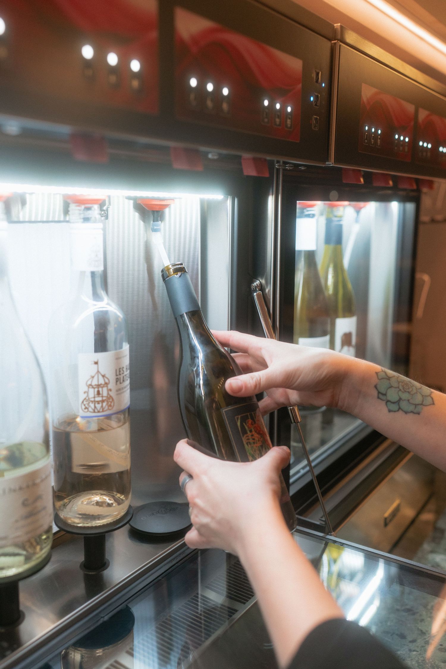 Tattooed hands retrieving a wine bottle from an illuminated wine preservation and dispensing machine.