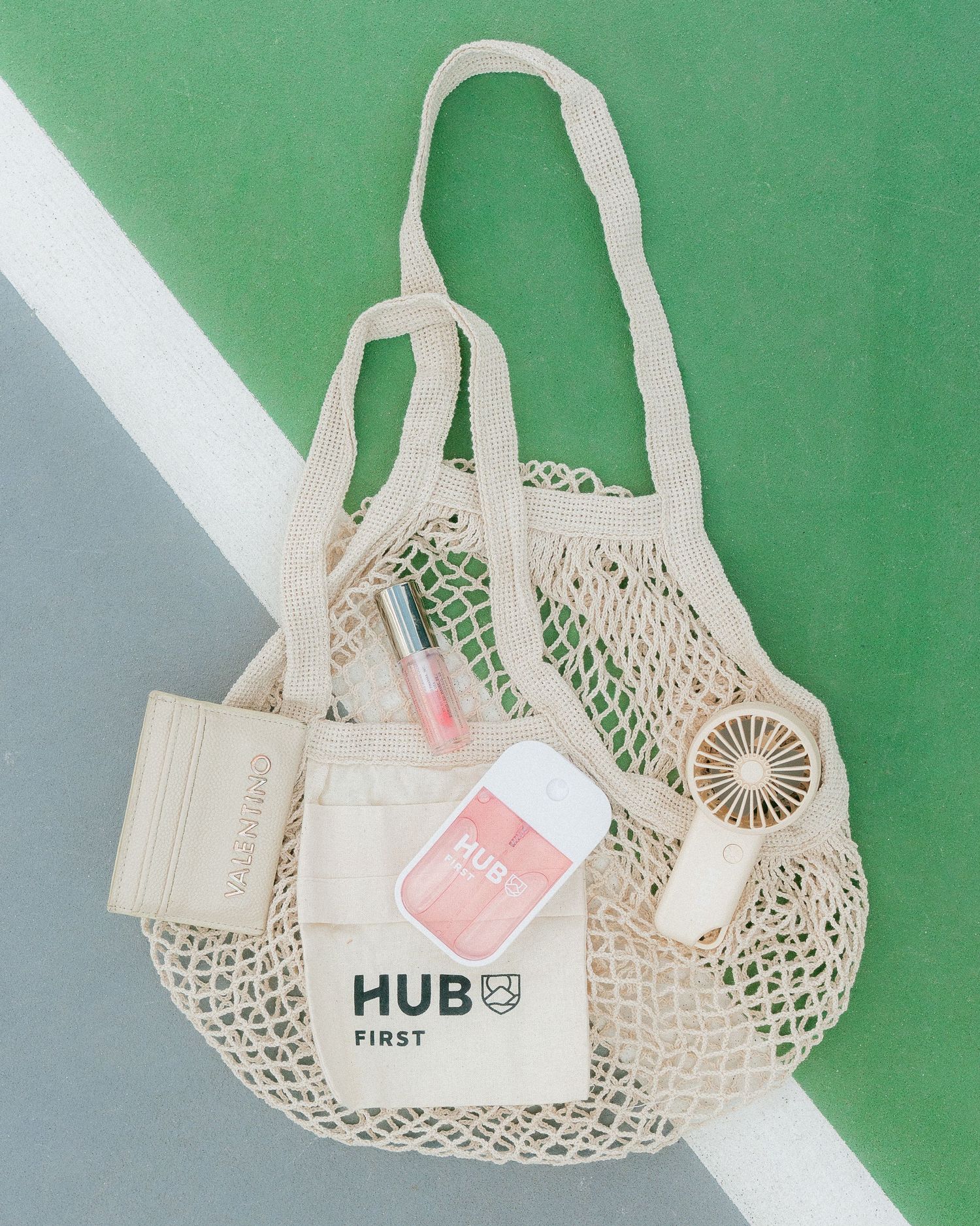 Hub First branded mesh tote bag with accessories on a green tennis court surface.