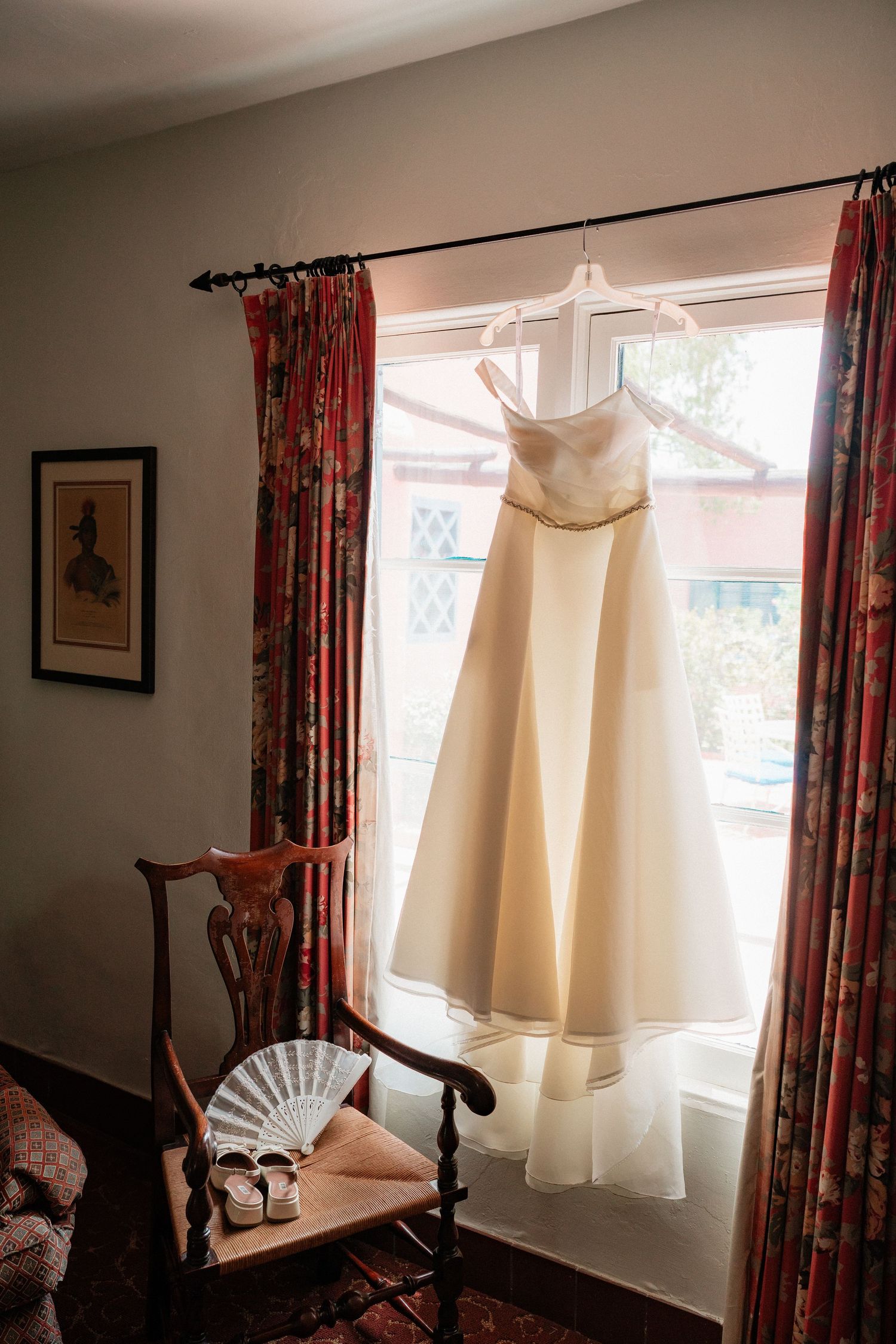 Strapless ivory wedding gown hanging in window with lace fan and bridal shoes on wooden chair below.