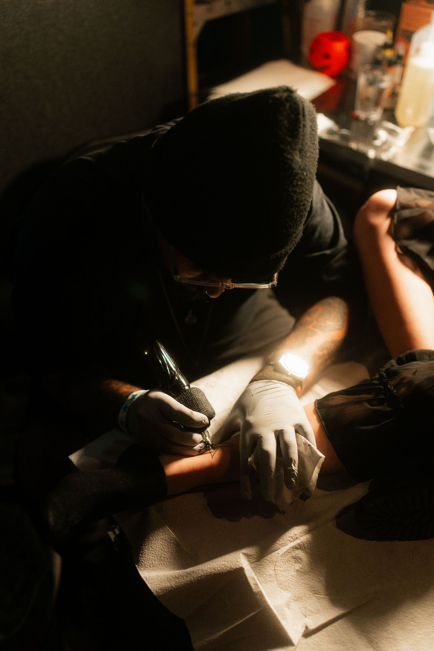 Tattoo artist in black wearing gloves carefully tattooing a client's arm under bright light.