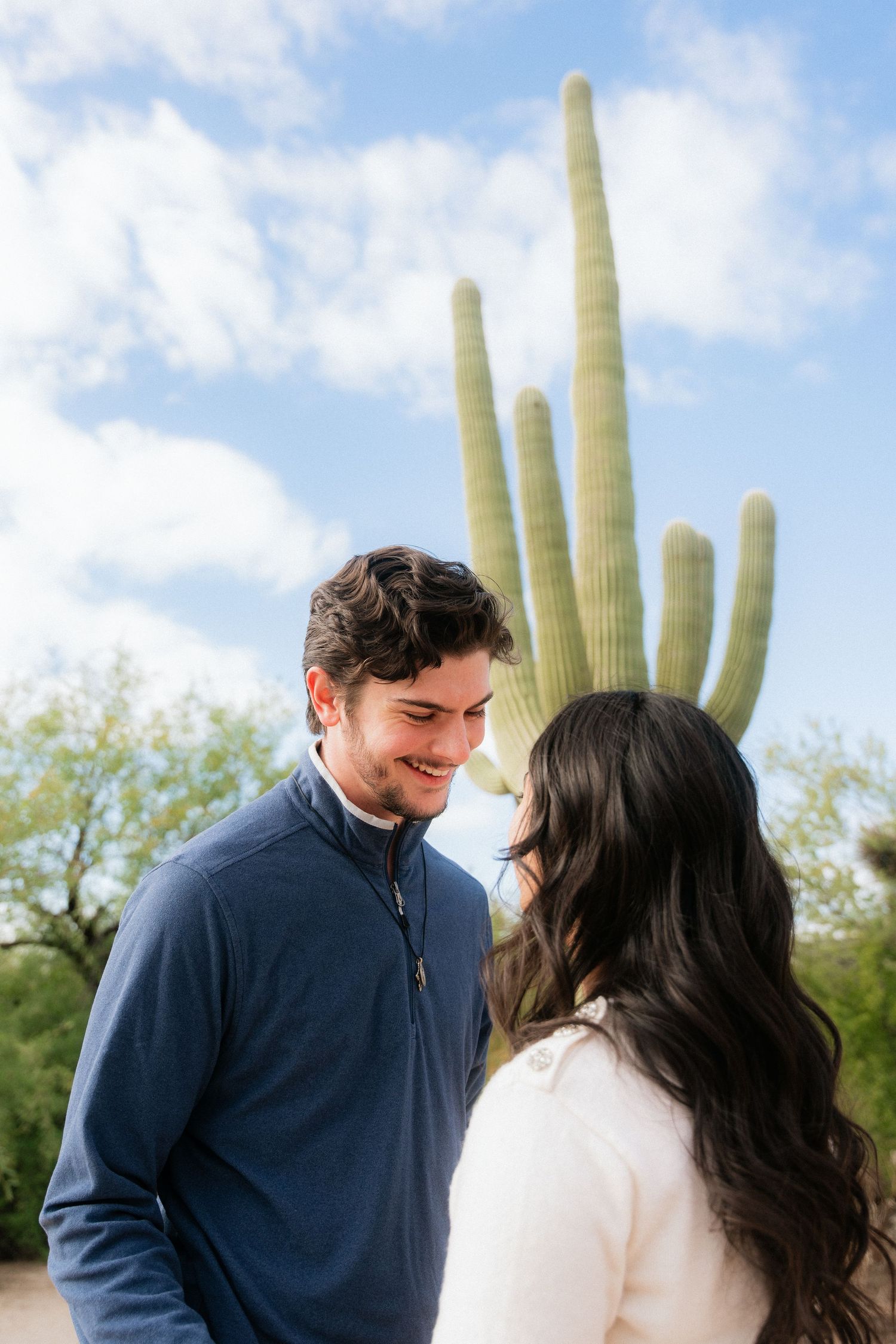 A smiling man gazes at a woman with a tall saguaro cactus and blue sky behind them.