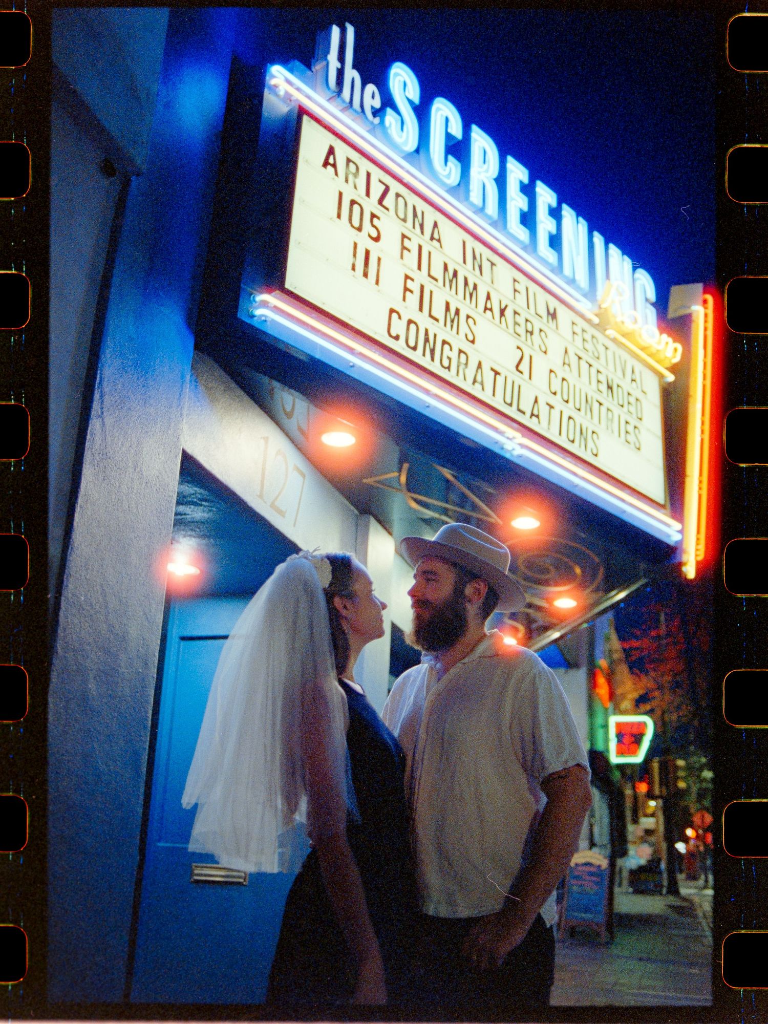Couple in wedding attire embracing under The Screening Room neon sign at night in Tucson, Arizona.