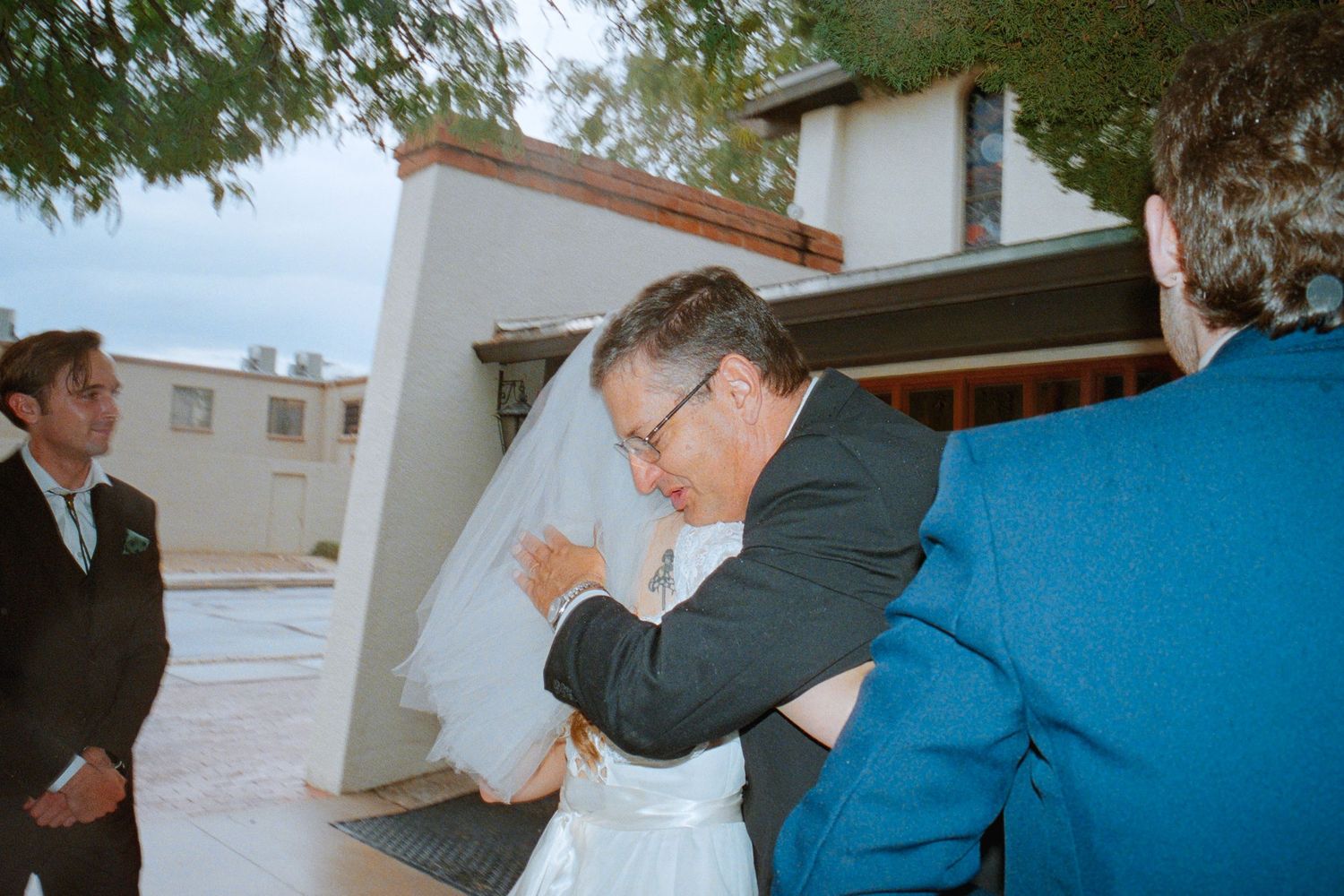 Father embracing bride in white wedding dress outside venue before ceremony.