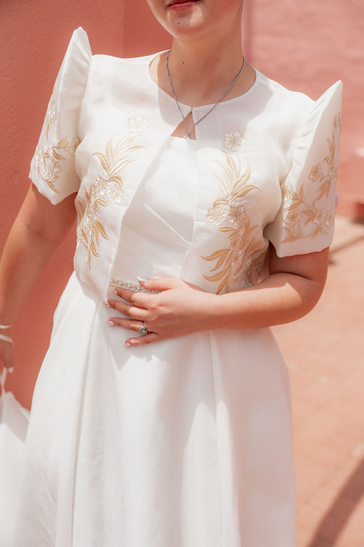 Bride in white embroidered dress with puff sleeves and gold floral details, wearing rings and a delicate necklace.