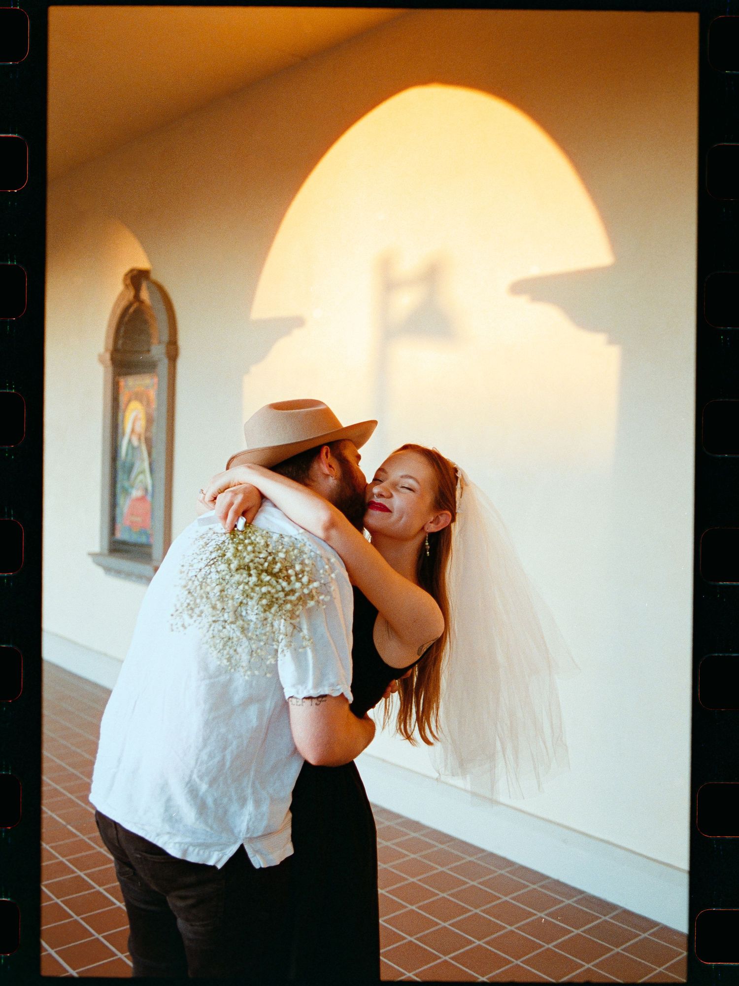 Joyful bride in black dress and veil embracing groom in hat inside sunlit church corridor.