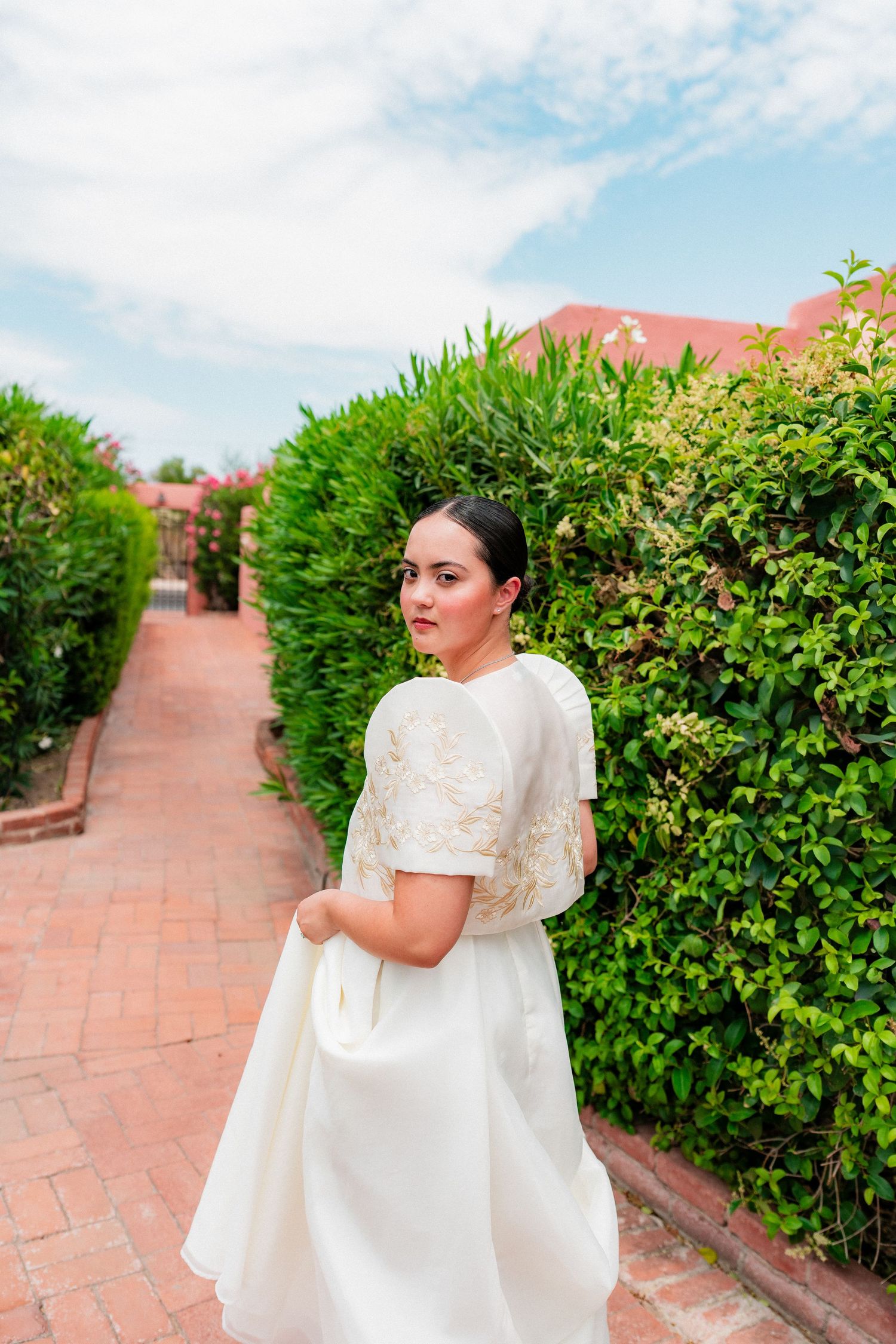 Bride in white embroidered Filipino Barong gown walks along garden path lined with lush green hedges.