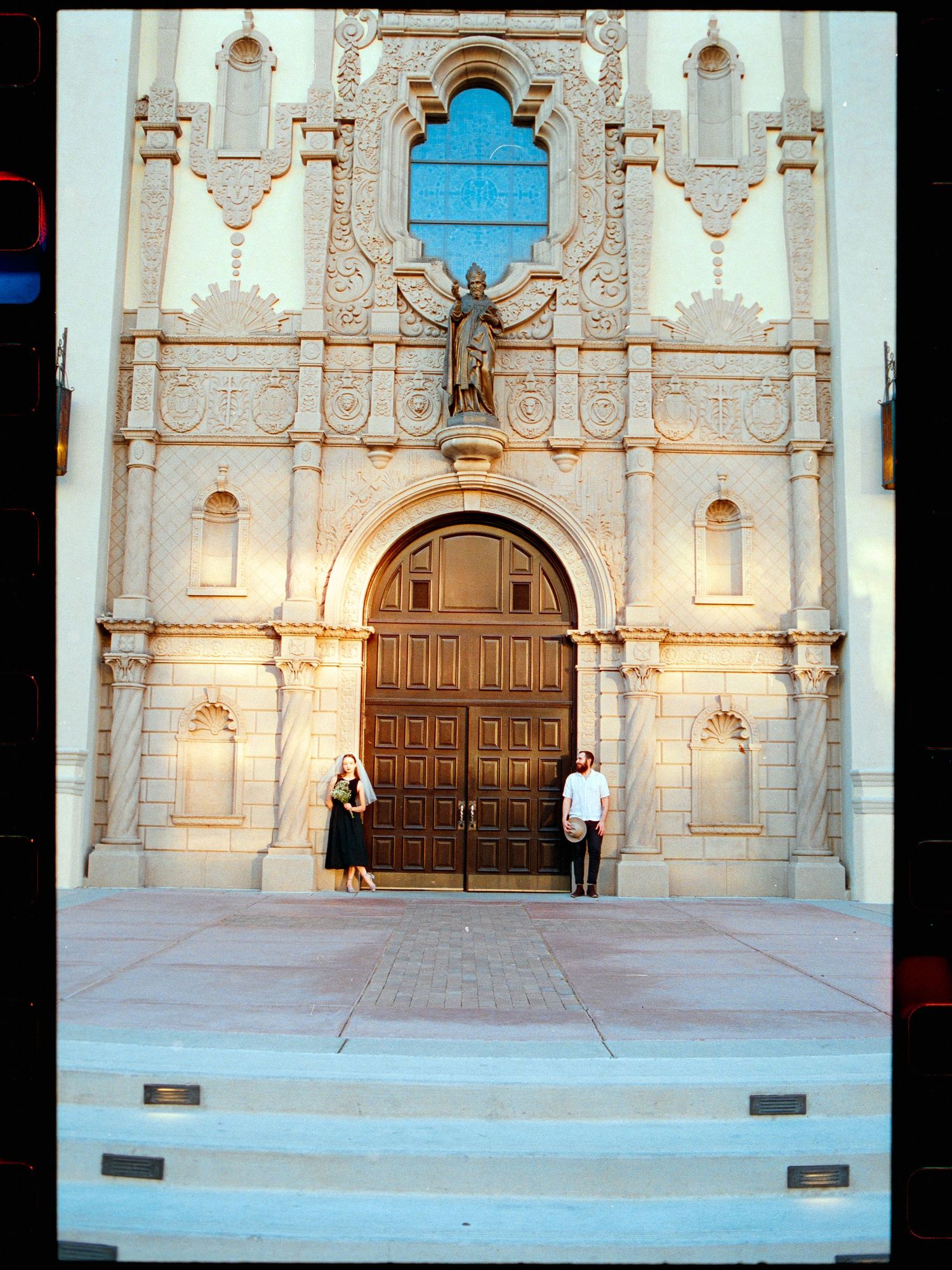 Wedding couple standing before ornate Spanish Colonial Revival church entrance with large wooden doors.
