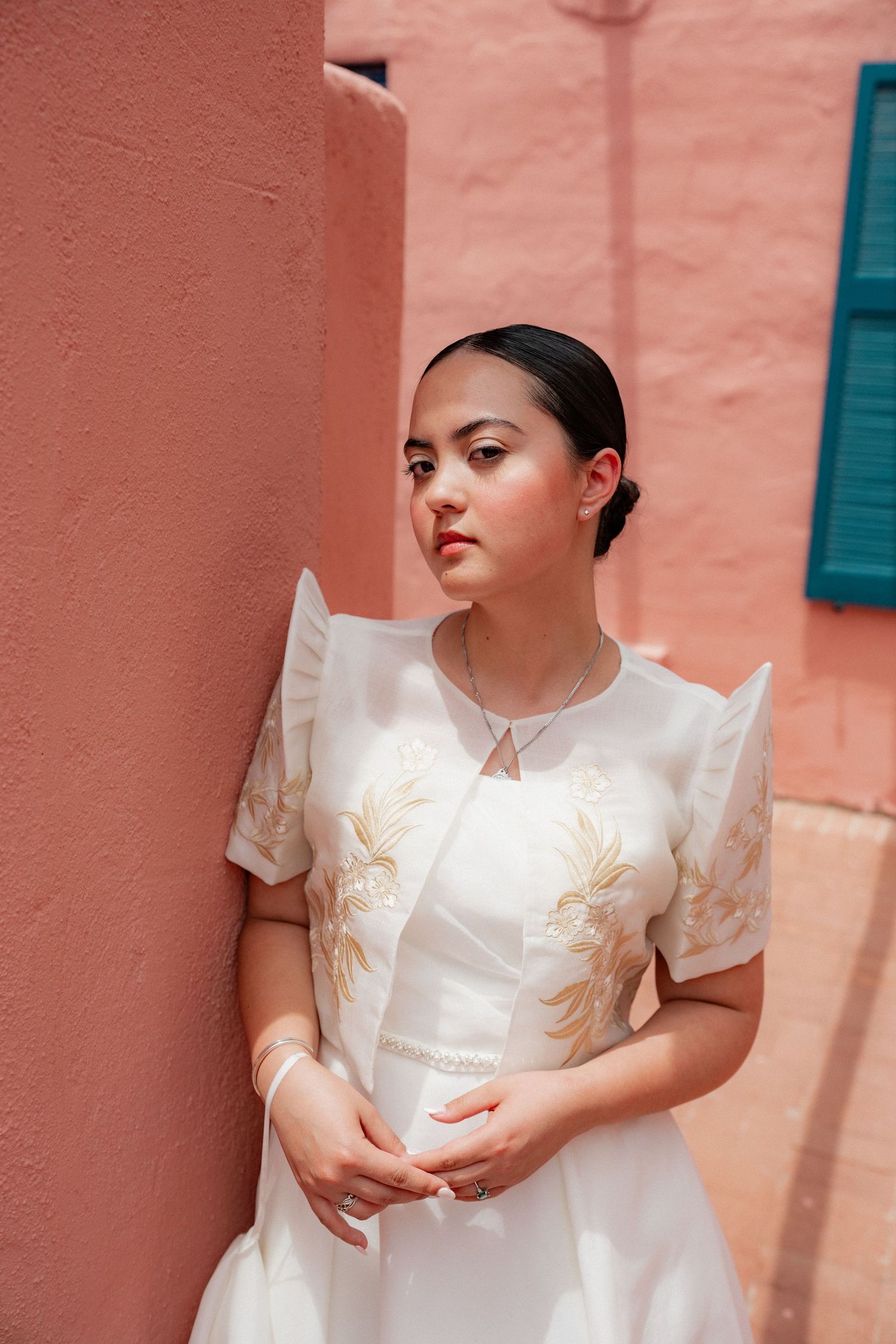 Bride in white embroidered Filipino Barong wedding dress leans against pink wall with teal shutters behind.