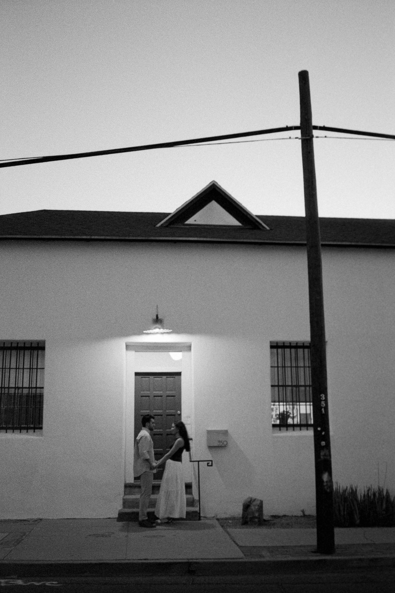Black and white photo of a couple holding hands outside a white building at dusk.