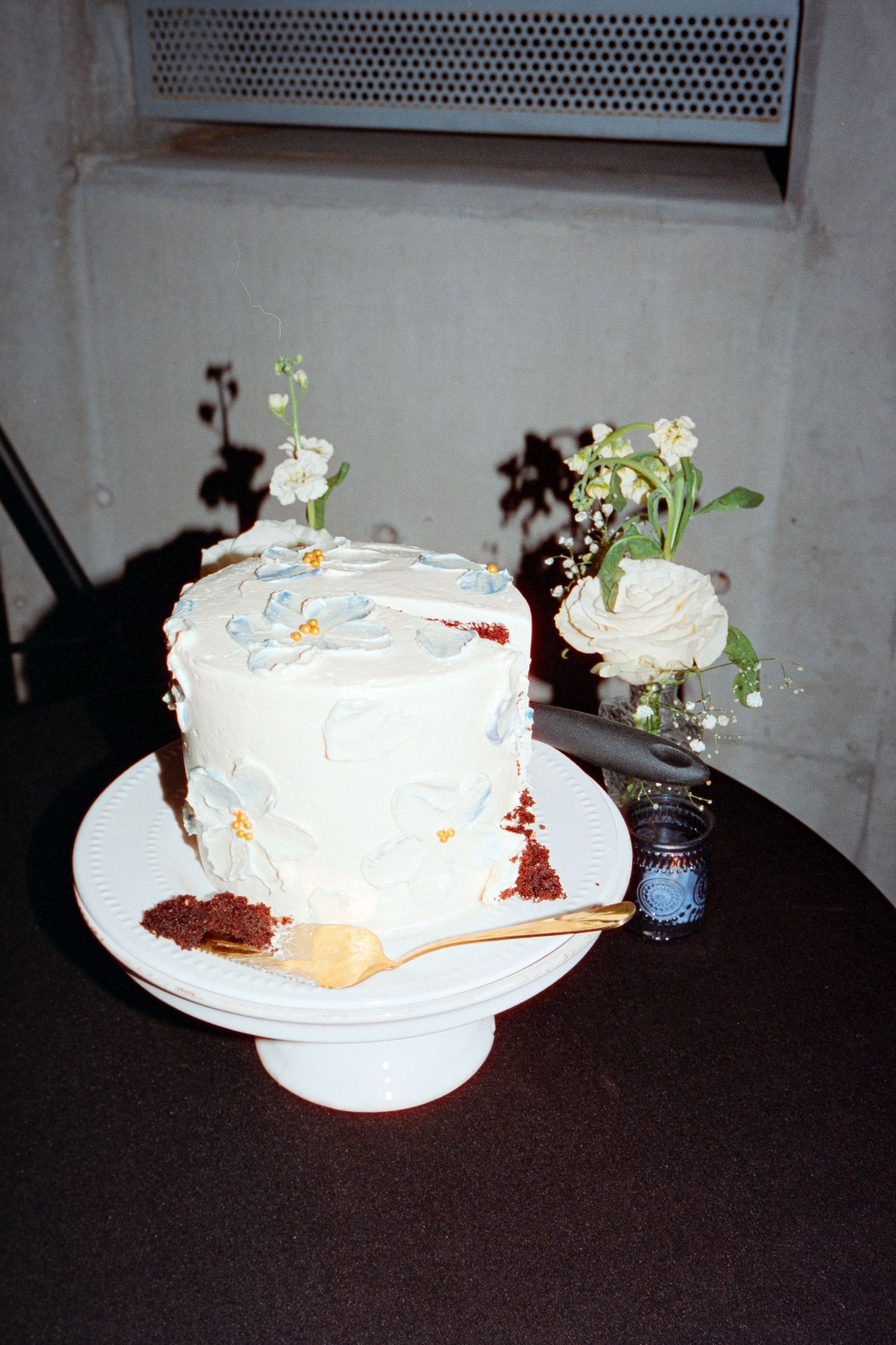 Elegant white wedding cake with blue floral decorations on a pedestal stand beside white flowers.
