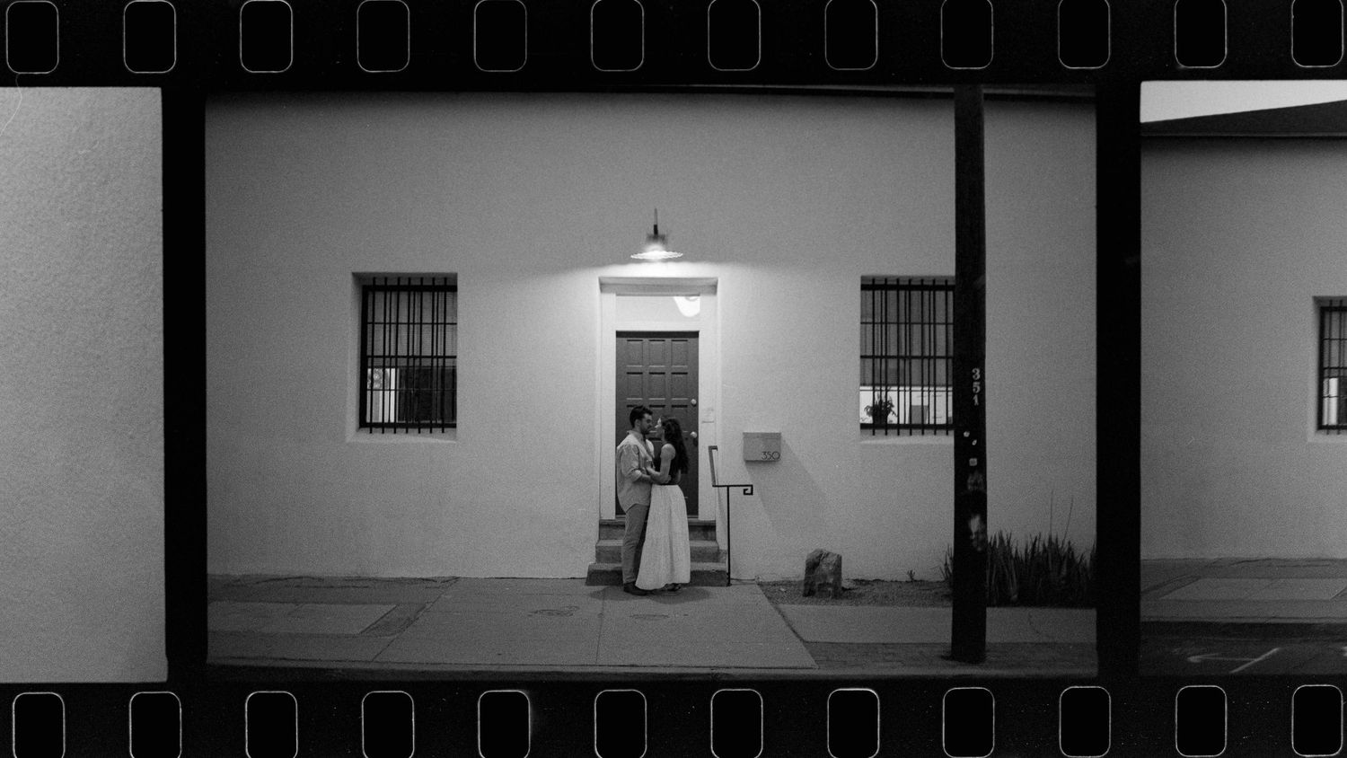 Black and white film strip photo of couple kissing under entrance light on quiet night street.