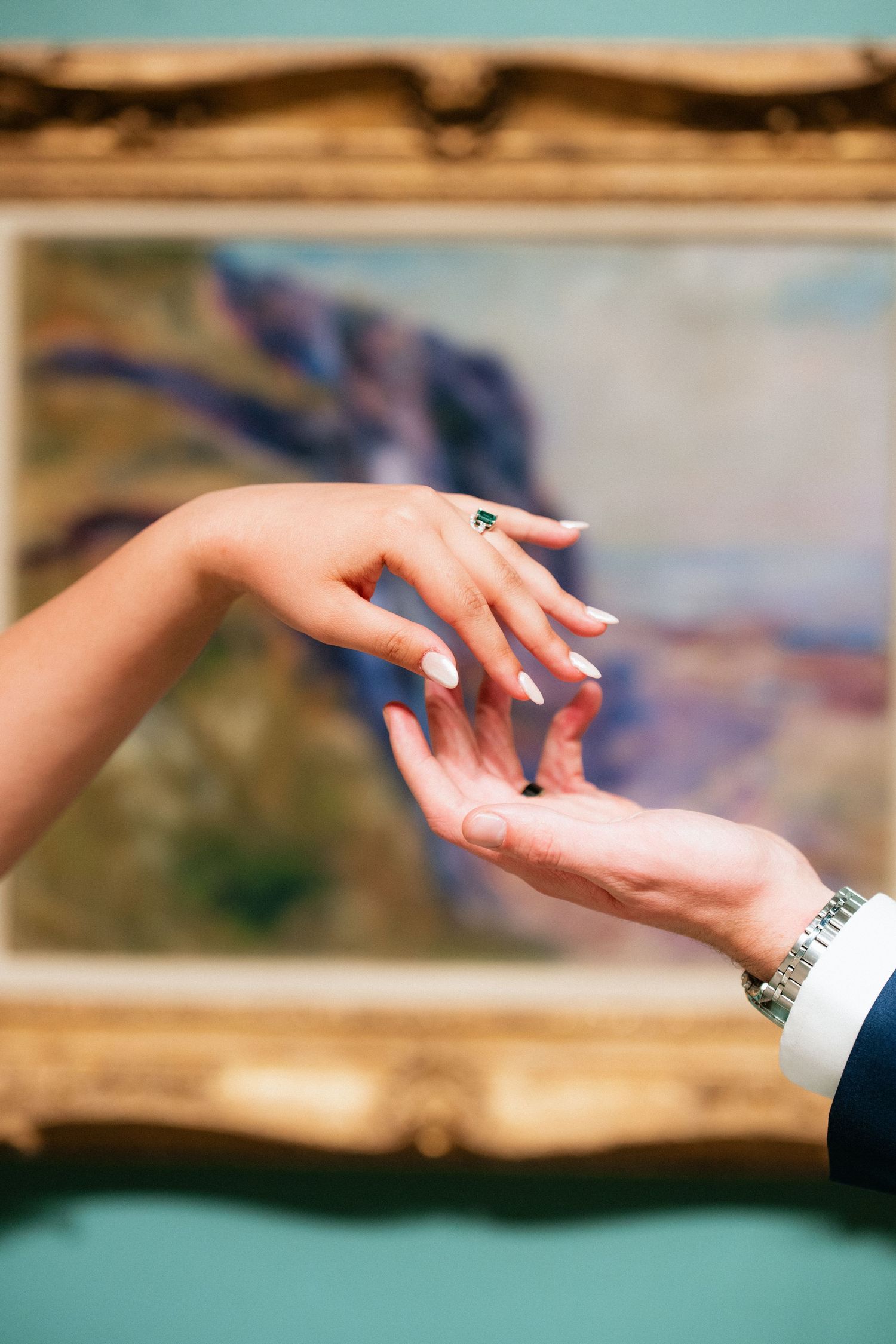 Close-up of bride and groom holding hands, showcasing emerald engagement ring and wedding bands in art museum.