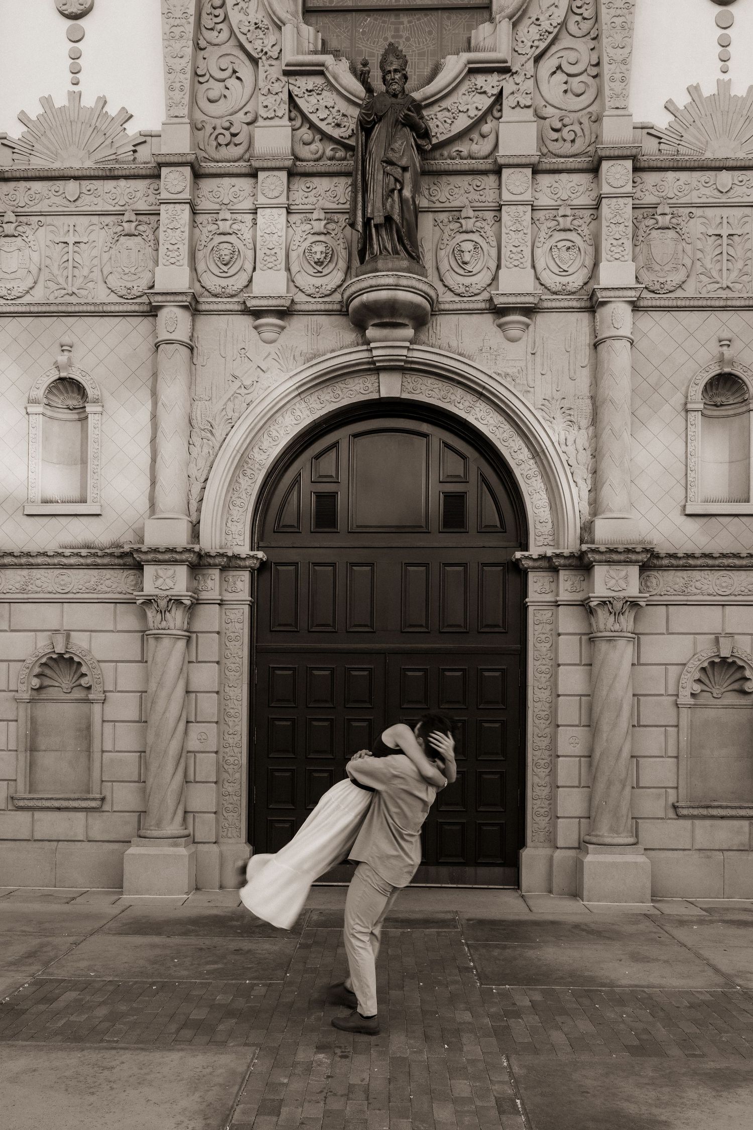 Black and white photo of couple embracing in front of an ornate church arched doorway.